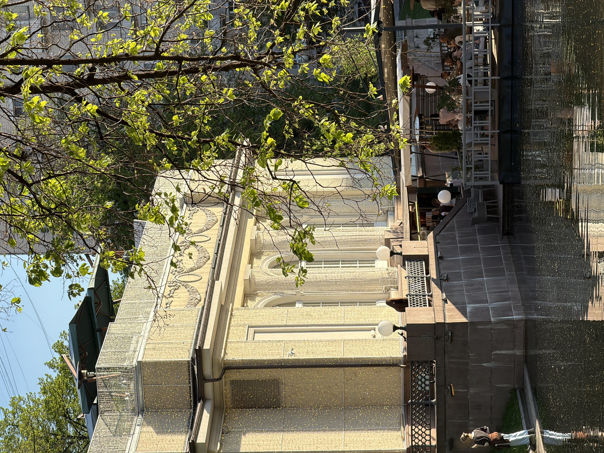 The image captures a sunny outdoor scene in Moscow, Russia, near a body of water, likely a canal or river. The architecture is characterized by a light-colored, multi-story building with ornate facade details, including arched windows and decorative molding. Tiny yellow lights are installed on the outside of the building. Branches with fresh green leaves frame the top of the image, suggesting it's springtime.
To the right of the building, there is a patio area with tables and chairs, seemingly part of a restaurant or cafe. People are seated at some of the tables. A pedestrian is seen near the water's edge on the lower right. The water reflects the building and the patio, creating a distorted mirroring effect. The sky is visible in the upper left corner and appears clear.
