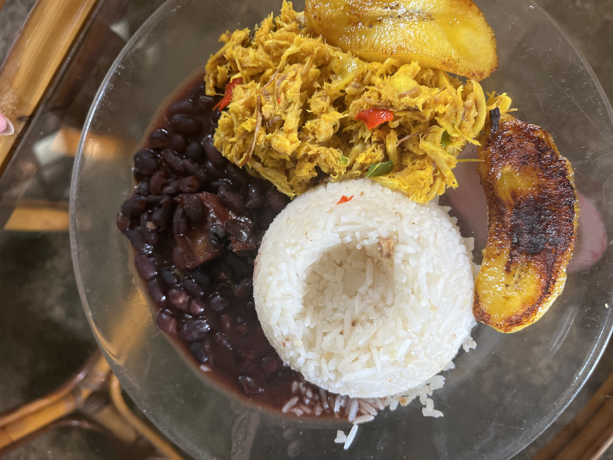 A close-up, overhead shot shows a plate of food on a glass surface, with a blurred background that appears to be a wooden table and chair. The plate contains a portion of white rice, a pile of shredded yellow meat, and a serving of dark beans. There are also two slices of fried plantain, one partially visible on the right and another whole slice at the bottom. The shredded meat has red chili peppers mixed in. The overall impression is of a home-cooked meal. The scene is devoid of any explicit indications linking it to Juan Griego, Venezuela.