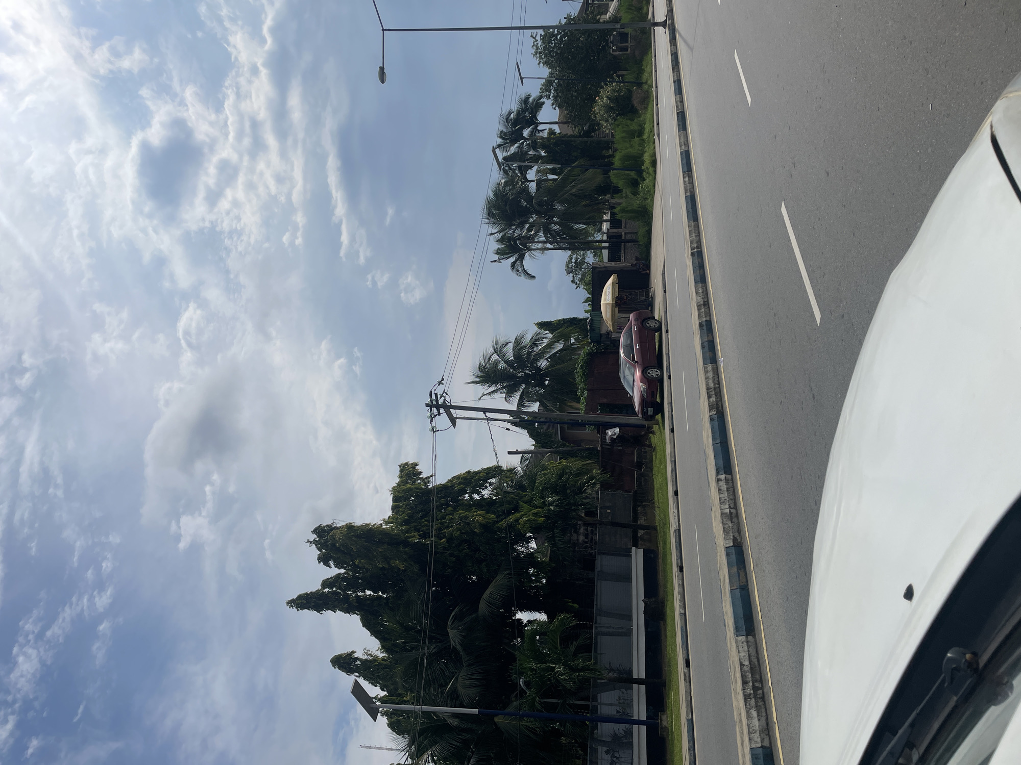 The image presents a daytime view from within a white vehicle, looking across a road towards a tree-lined roadside. The perspective is from the front right side of the car, with its white hood and a portion of the windshield visible in the foreground.
The sky, dominating the upper part of the frame, is a clear blue with scattered, fluffy white clouds, suggesting bright, fair weather. A tall streetlight extends from the left side of the frame into the sky.
The road is a multi-lane asphalt surface, likely a main thoroughfare. White dashed lines mark the lanes, and a concrete-curbed median strip separates the traffic directions, featuring some green grass.
Beyond the road and median, a dense strip of vegetation lines the street. Numerous tall palm trees with distinctive fronds are prominent, interspersed with other leafy green trees, creating a lush, tropical appearance. Utility poles with overhead power lines are visible among the trees.
A red sedan is parked or stopped on the side of the road, partially obscured by the trees. Next to it, an open yellow umbrella provides shade, hinting at a roadside vendor or a resting spot. Another streetlight can be seen further down the road amidst the greenery.
The scene portrays a typical urban or suburban environment. The abundant palm trees and the general aesthetic are consistent with the tropical climate of Port-Harcourt, Nigeria. No individuals are visibly present, nor is any legible text apparent. The atmosphere is calm and ordinary, capturing a moment from a drive.