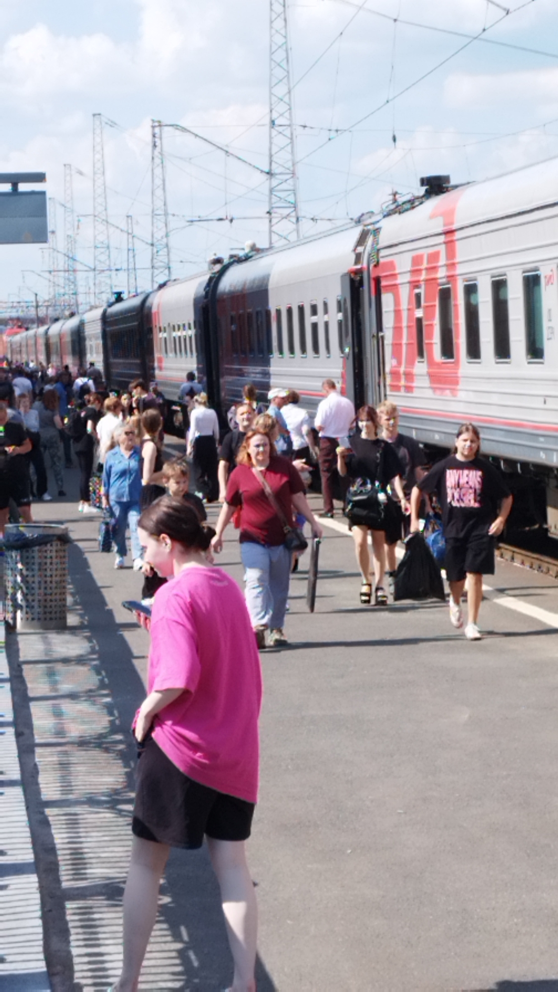 The image shows a busy outdoor scene at a train station, likely in Oryol, Russia, given the context. A long train with red and silver cars is parked at the platform, with its doors open. Numerous people, dressed in casual summer clothing, are walking on the platform, some in groups, others individually. Some appear to be disembarking from or boarding the train, while others are simply passing through. The platform is paved and has some trash receptacles. Overhead, power lines and supporting structures are visible, typical of a railway environment. The sky is partly cloudy. A young woman in a bright pink t-shirt and black shorts is in the foreground, facing away from the camera and looking at her phone.