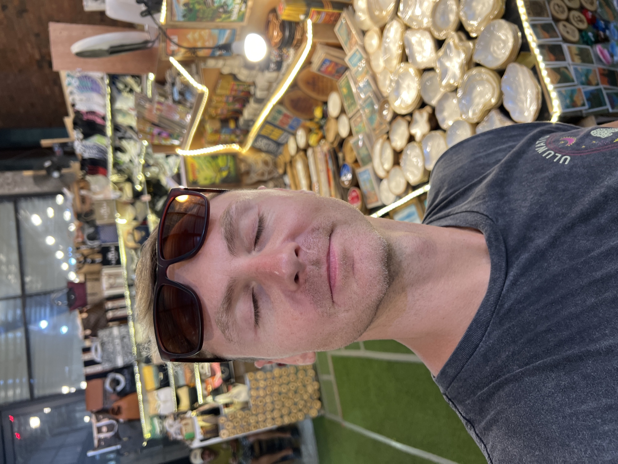 A man with short blond hair, wearing sunglasses and a gray T-shirt with the word "ULUWATU" and graphics, stands in what appears to be a retail shop with numerous goods on display. The shop seems to be located in Kuta Selatan, Indonesia, as inferred from the writing on the shirt. The man's eyes are closed. Overhead and in the background, shelves and displays hold a variety of items including rounded, possibly decorative objects, as well as other merchandise, illuminated by lights. The surroundings have an informal, market-like atmosphere.