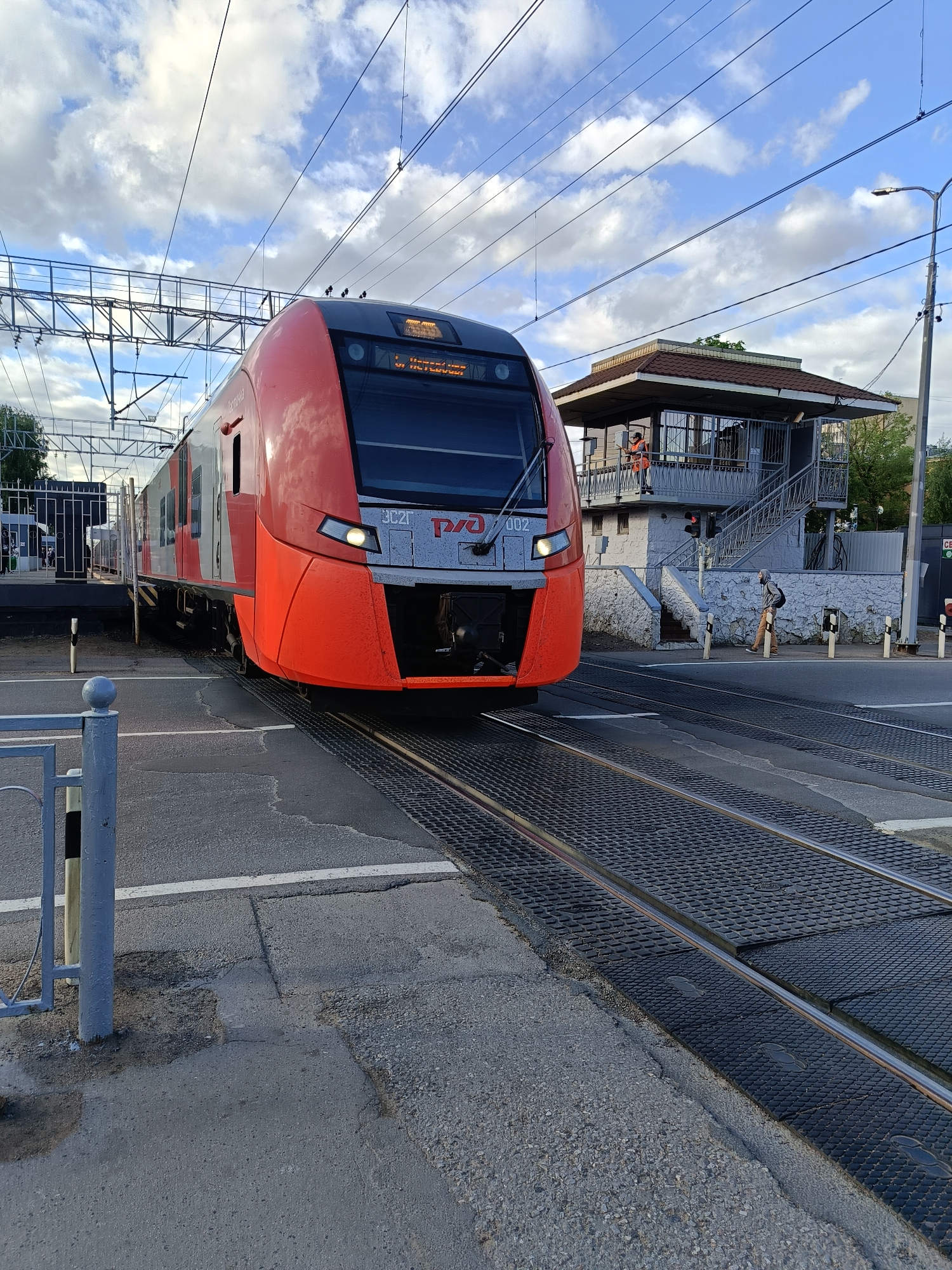 The image depicts a modern red and gray electric train crossing a road in Saint Petersburg, Russia. The train bears the Russian Railways logo ("РЖД") and is approaching a railway crossing with a small, two-story control tower to the right. A railway worker in orange is visible in the tower. A pedestrian is walking near the crossing. The crossing is equipped with safety features, including barriers and signal lights. The sky is partly cloudy.