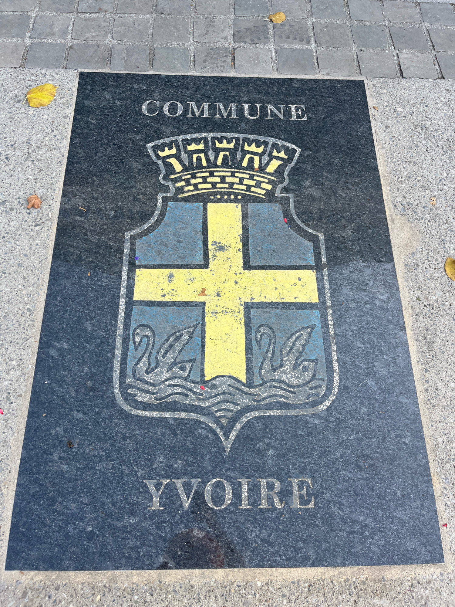 The image shows a close-up, eye-level view of a black granite slab embedded in the pavement of a European city. The slab features a coat of arms and text. The coat of arms is centrally located, with a yellow cross on a blue background, and two white swans below the cross, with wavy lines representing water. Above the shield is a yellow crown with three points. The text "COMMUNE" is inscribed in silver capital letters above the coat of arms, and "YVOIRE" is inscribed in similar letters below. The surrounding pavement consists of small, grey cobblestones. A few yellow and brown fallen leaves are scattered on the pavement and the granite slab. The lighting suggests it is daytime. The inscription "YVOIRE" indicates the location is the city of Yvoire, France.