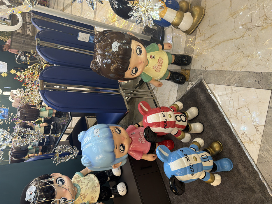 A high-angle shot displays a collection of cartoonish figurines on a glossy surface and a grey rug. The figurines include a girl with brown hair, another with blue hair, a red and white "8" figure, and a blue and white "10" figure. Some figures have text that reads "wang dao jia ju". Blue chairs with chrome frames are nearby. A display of decorative lights and items on a mirrored surface is visible in the background. The scene is brightly lit and appears to be inside a retail or showroom environment.