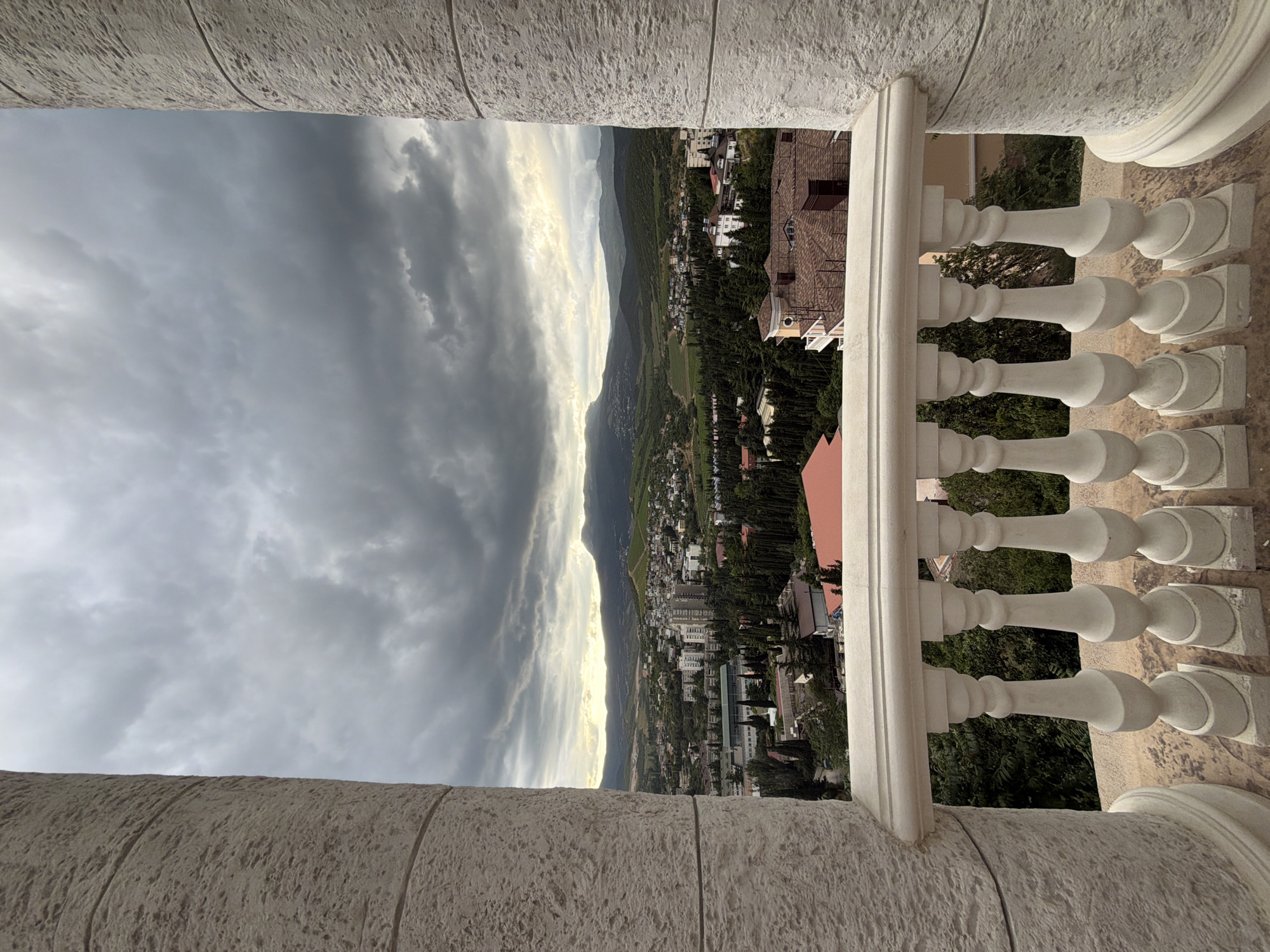 A high-angle shot captures a panoramic view of Alushta, Ukraine, under a dramatic, overcast sky. The foreground features a white balustrade and parts of two textured white columns, framing the cityscape. Buildings, varying in height and architectural style, are nestled amongst dense evergreen trees. Beyond the immediate urban sprawl, rolling hills and mountains stretch towards the horizon. The light suggests either early morning or late afternoon, with a subdued brightness breaking through the clouds. No people are visible in the image.
