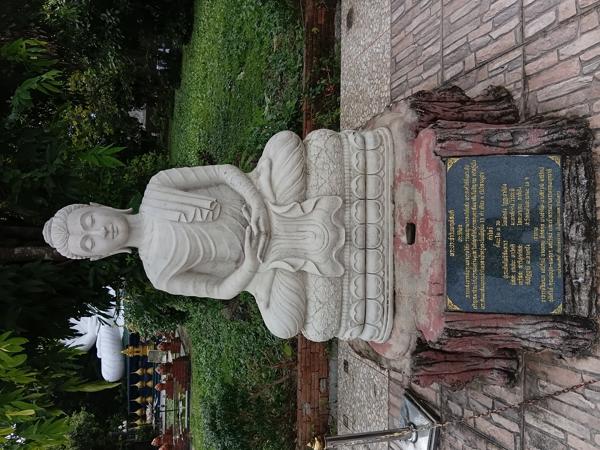 The image shows a statue of a seated Buddha in a garden setting. The statue is made of light-colored stone and is intricately carved, depicting the Buddha in a meditative pose. It rests on a decorative base, which is supported by a rough, wood-like pedestal. In front of the pedestal is a dark plaque with Thai script, likely providing information about the statue.
The background features lush green foliage and trees, suggesting an outdoor, possibly temple or park environment. In the distance, partially obscured by leaves, is another, larger white statue of a reclining Buddha. The ground around the statue is paved with interlocking stones. A metal railing with a golden top is visible to the left of the Buddha statue. The overall scene is tranquil and appears to be a place of reverence. The provided information indicates that this location is in Ban Ruammit, Thailand.