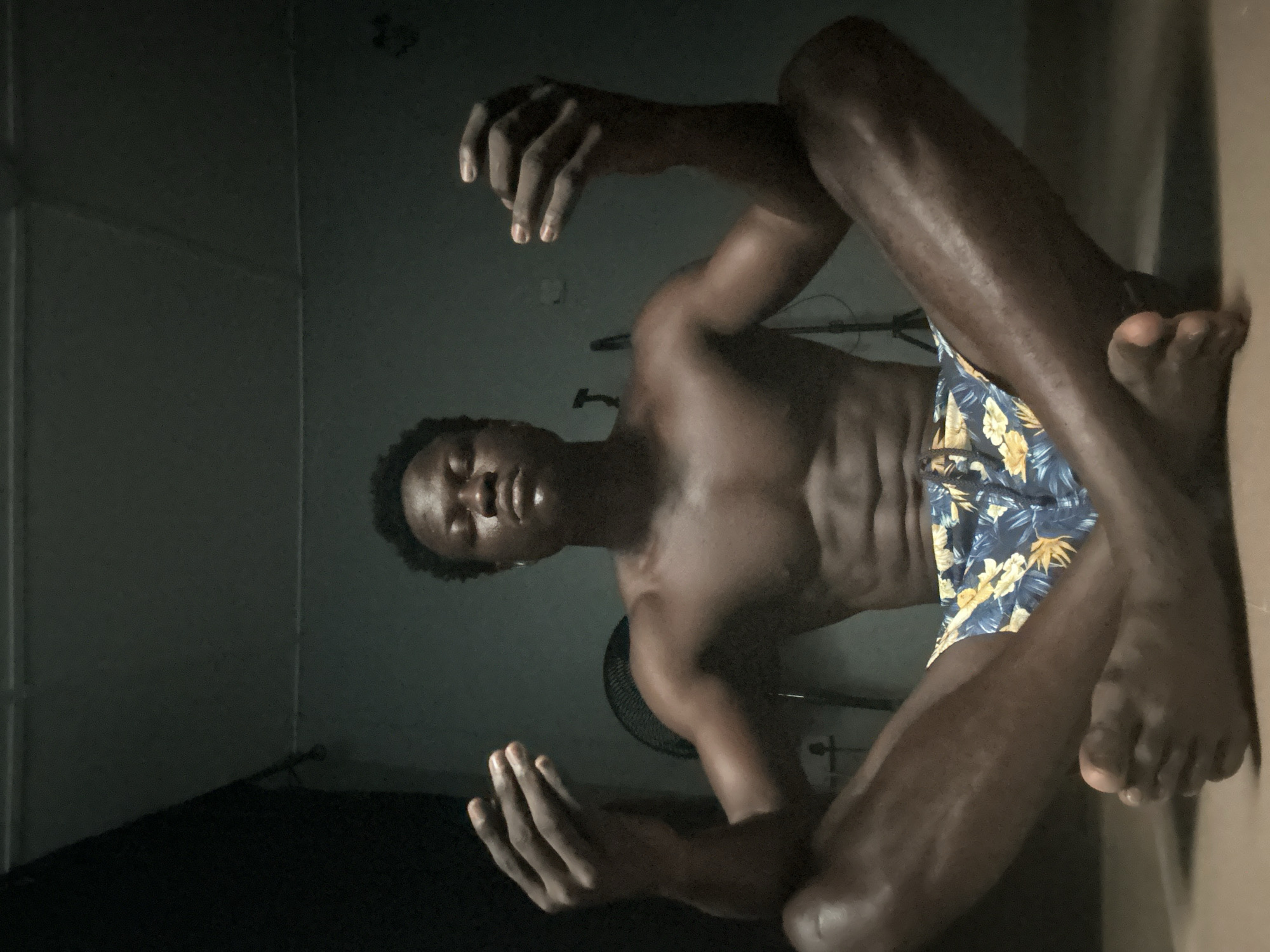 A young Black male is pictured from a low angle, sitting with his legs outstretched and bent at the knees. He is shirtless, wearing floral-patterned swim trunks. His body is glistening, suggesting he may have recently showered or is in a humid environment. His head is tilted back, eyes closed, and his face shows a relaxed or meditative expression. His hands are raised and open, palms facing upwards. In the background, a dark wall and a fan are visible. The lighting is dim, with some light casting shadows, suggesting it might be nighttime or an indoor setting with limited light. The image appears to be taken in Badagry, Nigeria, as indicated by the location context. There is no visible text in the image.