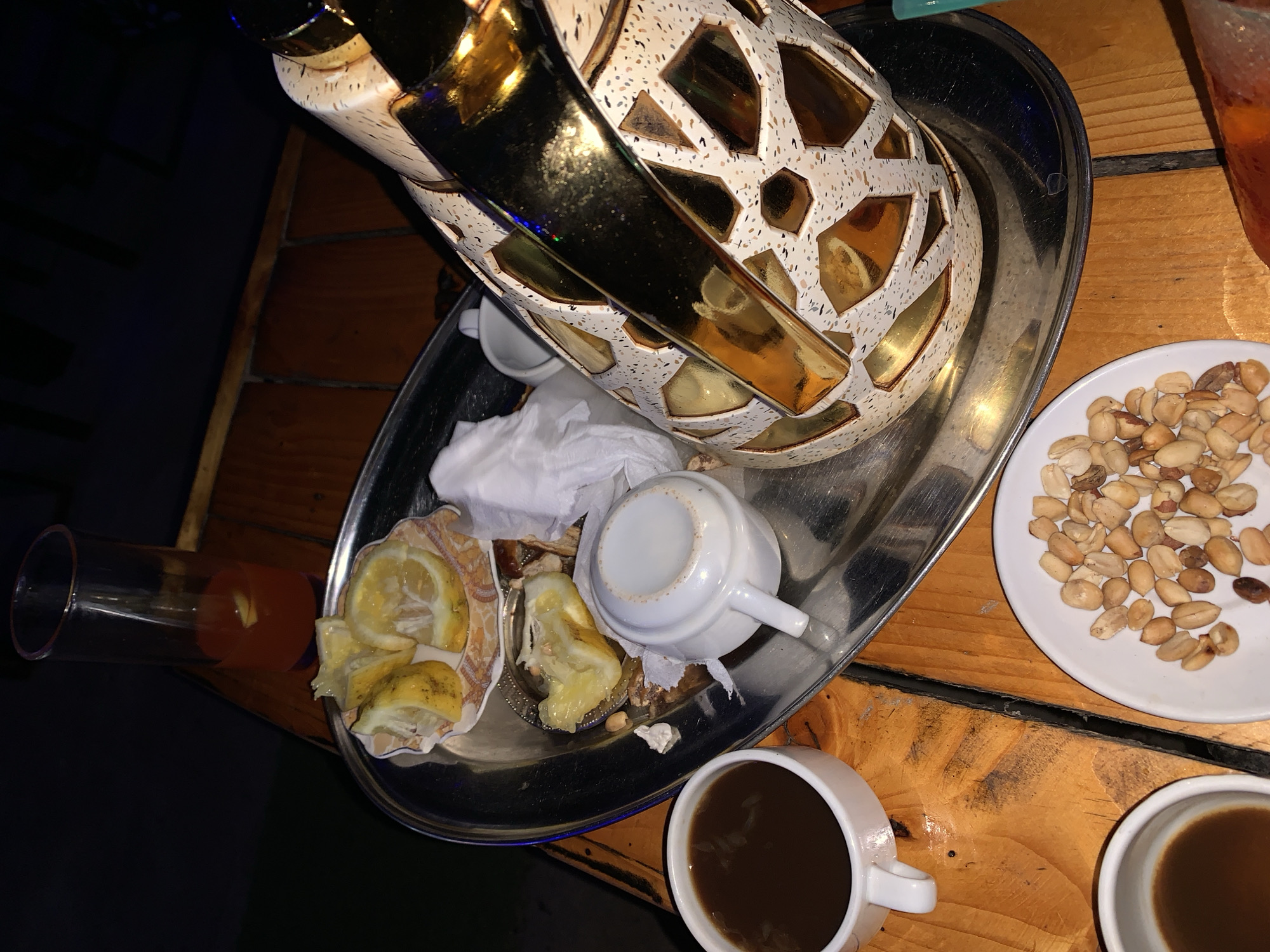 An overhead shot captures a wooden table laden with a tray of refreshments. On the tray, a decorative thermos with gold accents sits next to a small white creamer and a stack of used napkins. Several wedges of lemon are also present, some appearing partially consumed. To the left of the tray, two white cups of dark liquid, likely coffee, are visible, one slightly closer to the foreground. A plate of peanuts sits in the lower right quadrant of the image, and a tall glass filled with a red liquid, possibly juice, is positioned behind the tray. The scene is dimly lit, suggesting an evening setting.