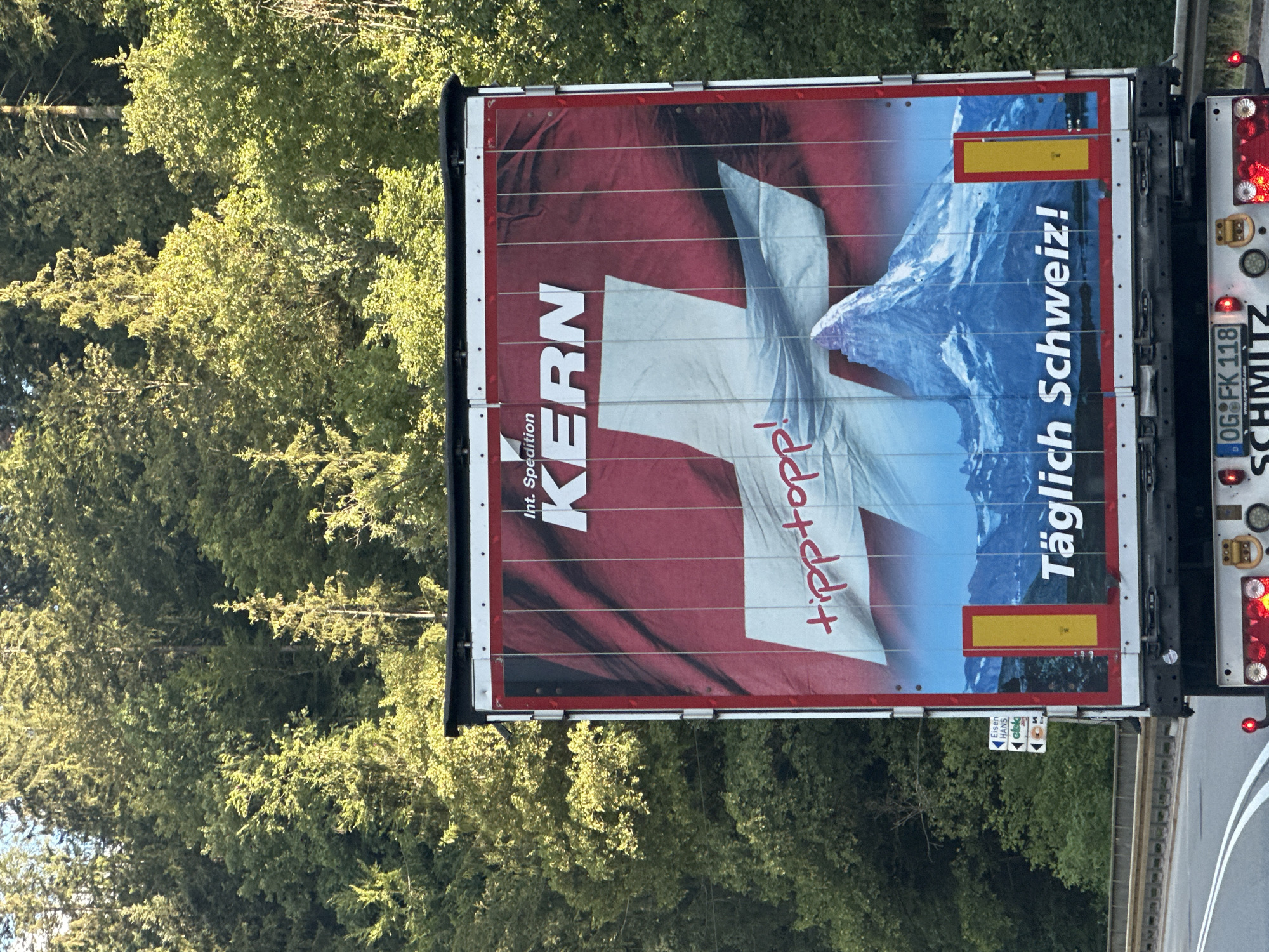 This image shows the rear of a truck trailer with a prominent advertisement. The trailer is red with white vertical stripes and features a large Swiss flag graphic. The words "Int. Spedition KERN" are printed in white at the top of the trailer, with "KERN" being significantly larger. Below this, the phrase "tipp topp!" is written in red script. In the lower portion of the trailer, a snow-capped mountain, resembling the Matterhorn, is depicted against a blue sky with a body of water and distant mountains. The German text "Täglich Schweiz!" is printed in white at the bottom. On the left side of the trailer, a small road sign with directional arrows and text like "Eisen Hans" is visible. The truck is on a road, with a dense forest of green trees forming the background. The scene appears to be in Schonach im Schwarzwald, Germany.