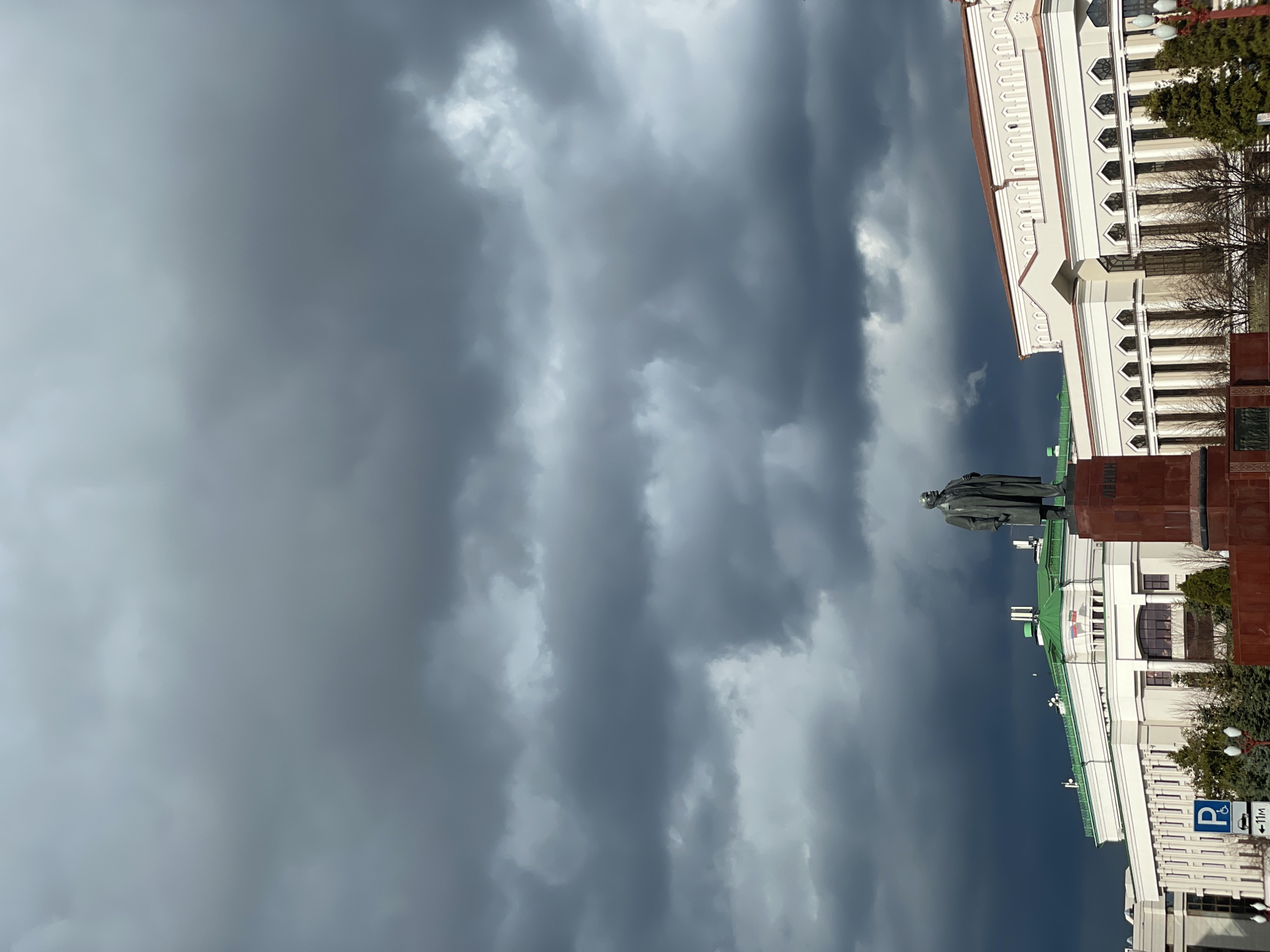 The image, correctly oriented, presents a monumental urban landscape dominated by a dramatic, overcast sky. Dark, heavy grey storm clouds fill the upper and central portions of the frame, with lighter patches indicating variations in cloud density.
In the foreground, a large, dark-colored statue of a man, likely Vladimir Lenin given the context of Kazan, Russia, stands on a red-brown stone pedestal. The figure is depicted wearing what appears to be a long overcoat.
To the right, a grand, multi-story building with striking white facades and elaborate architectural details occupies a significant part of the scene. The building features rows of decorative windows, some arched or hexagonal, and ornate molding along its structure. A section of the building exhibits a bright green roof or trim. In front of and around the building, a few bare trees and evergreen bushes are visible, suggesting a time of year outside of full summer foliage.
At the bottom right of the frame, a blue parking sign is clearly visible. It displays a white 'P' symbol and a wheelchair accessibility icon. Below this, a smaller white sign indicates "<- 11m", likely denoting a distance or direction for the parking facility.
The overall lighting is diffused and dim, consistent with heavily cloudy or pre-storm weather conditions. The scene is static, with no discernible human activity. The combination of historical architecture and the dramatic, brooding sky creates an imposing and somewhat solemn atmosphere.
