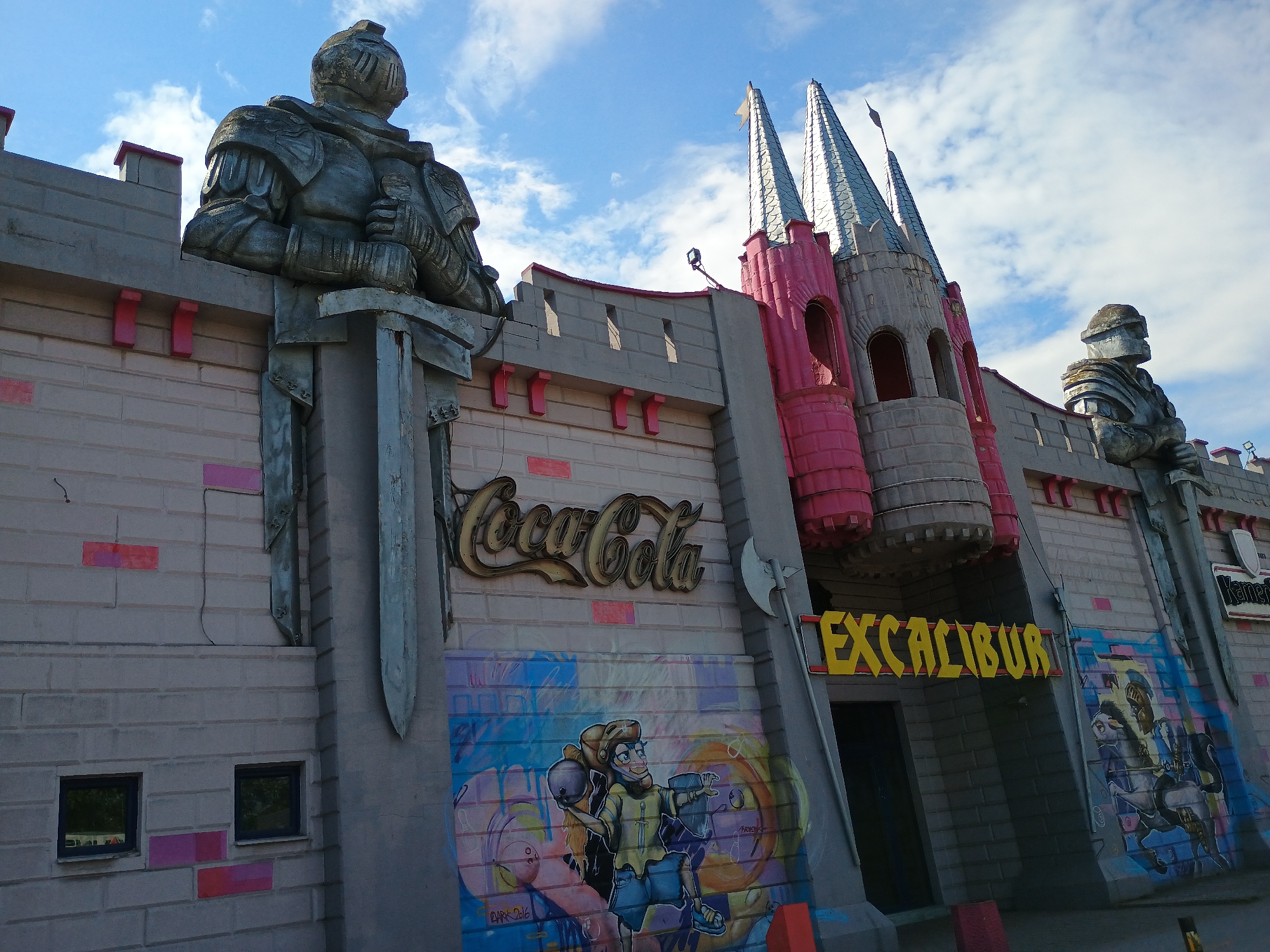 The image shows the facade of the Excalibur arcade in Unknown, Unknown. The building is designed to resemble a medieval castle, with two knight statues flanking the entrance. A large Coca-Cola sign is visible on the wall, along with graffiti art. The castle has pink and grey towers with silver roofs.