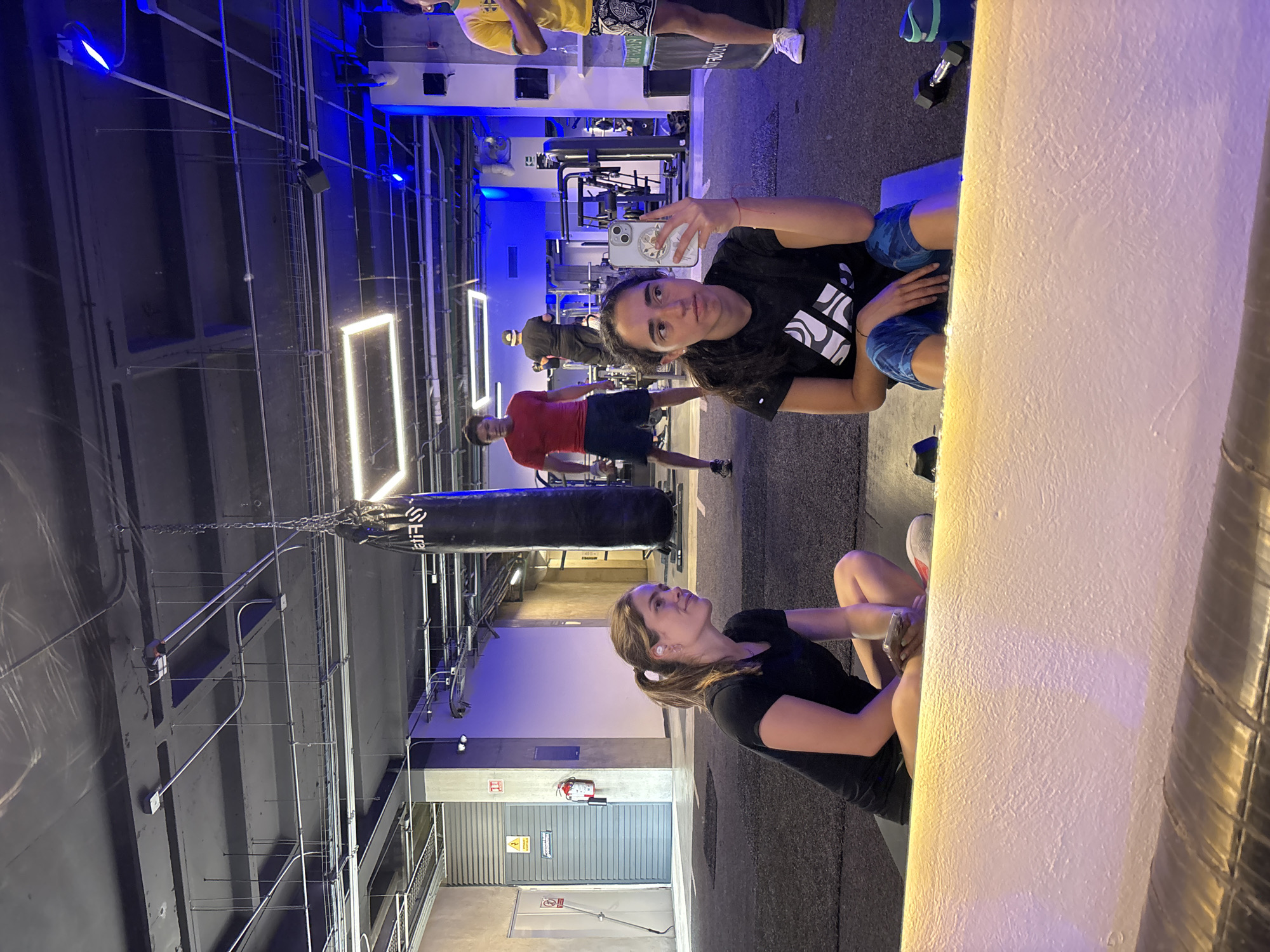 A selfie taken in a gym shows two women and several other people. One woman is taking the selfie with her phone, while the other woman is sitting on the floor in the reflection of a mirror. The gym has workout equipment and a punching bag. The ceiling is high and open, with metal structures and pipes visible. Blue and white lighting is used throughout the gym. The people in the background appear to be working out.