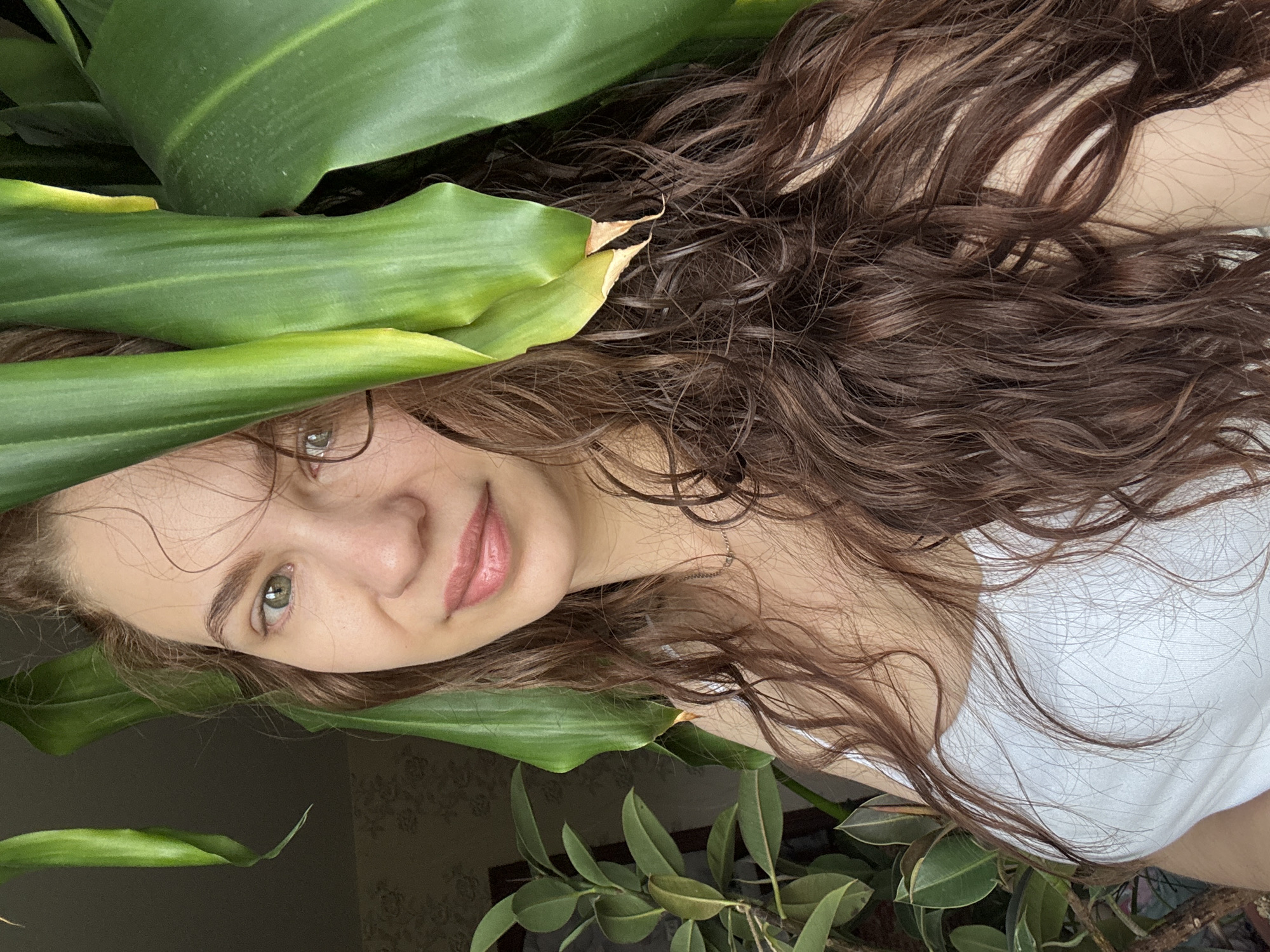 A young woman with light skin, long curly brown hair, and green eyes is partially obscured by the broad green leaves of a plant. She wears a white top and has a slight smile. The photo appears to be taken indoors, with more foliage visible in the background and potentially window. The location provided (Nizhnyaya Gar', Russia) is not verifiable from this image.