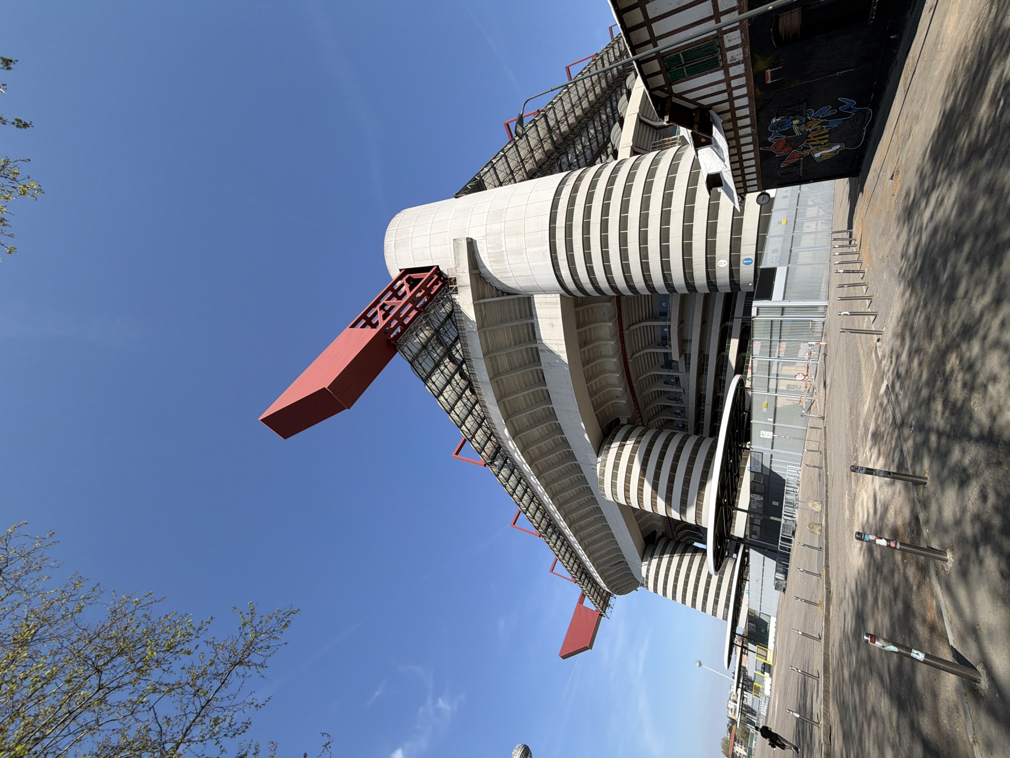 The image depicts a low-angle view of the San Siro stadium in Milan, Italy, under a clear blue sky. The stadium's external structure features cylindrical, white-and-black striped towers connected by a grid-like red steel support system. A section of concrete pavement and metal bollards is visible on the lower-right side of the frame. A tree branch appears in the lower-left corner. A pedestrian is present walking away from the stadium. Graffiti decorates the wall in the background.