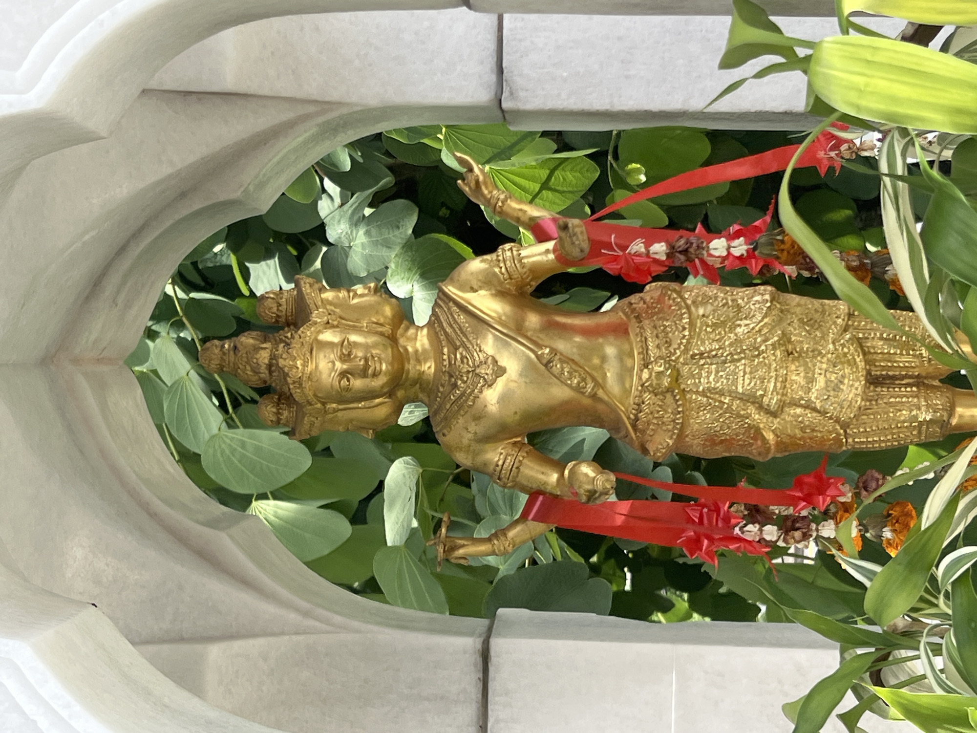 A golden statue of a multi-faced deity stands within a white arched alcove. The statue depicts a figure with four faces, each with serene expressions. The deity is adorned with intricate golden jewelry and clothing, including a crown with multiple tiers. Red ribbons and garlands with dried flowers are draped around the statue's arms and lower body. In the background, lush green foliage is visible through the arch. The statue is positioned outdoors, with green plants in the foreground. The architectural style of the alcove suggests it is part of a temple or shrine, potentially located in Pa Khlok, Thailand.
