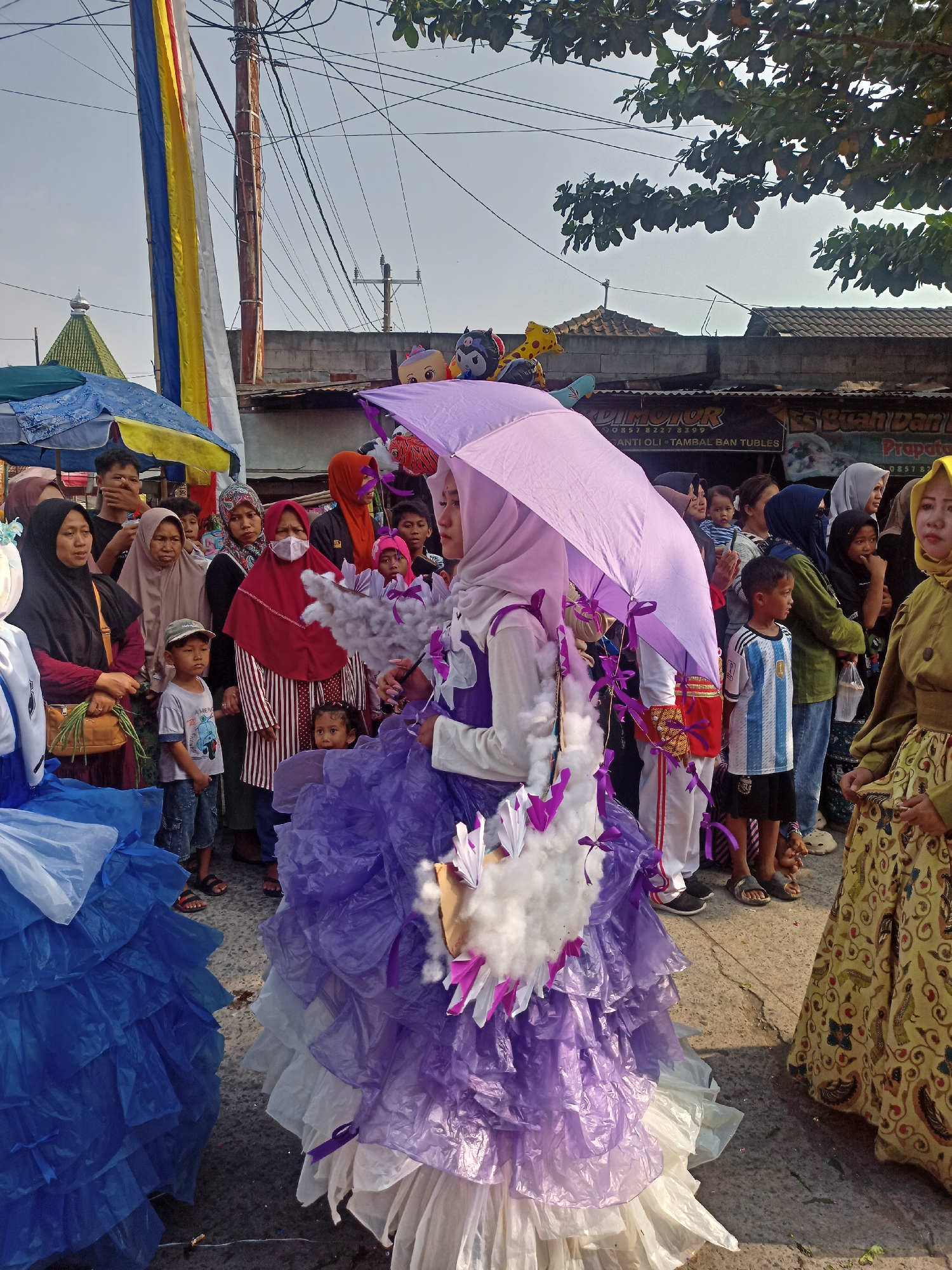 This is a photograph taken in Saren, Indonesia, depicting a festive outdoor event or parade. In the foreground, a young woman in a purple hijab and a voluminous costume made of layered purple and white material, resembling petals or ruffles, holds a large, light purple umbrella over her head. The costume is adorned with white, fluffy elements and purple accents. To her left, a portion of another costume, predominantly blue, is visible.
A crowd of people, mostly women and children, are gathered around, observing the parade. Many are wearing hijabs. The background reveals a street scene with buildings, possibly shops or houses, some with corrugated metal roofs. Overhead, there are power lines and a utility pole, along with tree branches. Balloons, some shaped like cartoon characters, are visible in the upper right portion of the image. Colorful flags, including a yellow, blue, and white striped one, hang from poles. The overall atmosphere appears to be lively and communal.