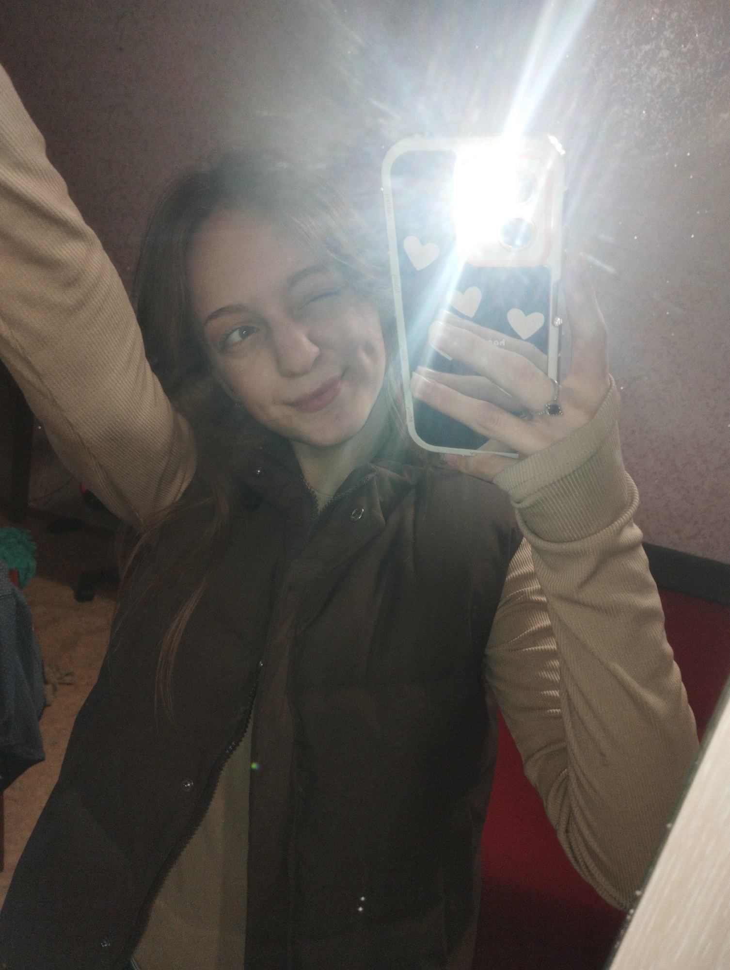 A young woman is taking a selfie in front of a mirror. She is wearing a brown vest over a tan long-sleeved shirt and has light brown hair. She is winking and has a slight smile. The phone she is holding has a white case decorated with hearts, and the flash is on, creating a bright light. The background appears to be a room with light-colored walls, but due to the angle and lighting, many details are obscured. There is no evidence to indicate that the image was taken in Saratov, Russia.