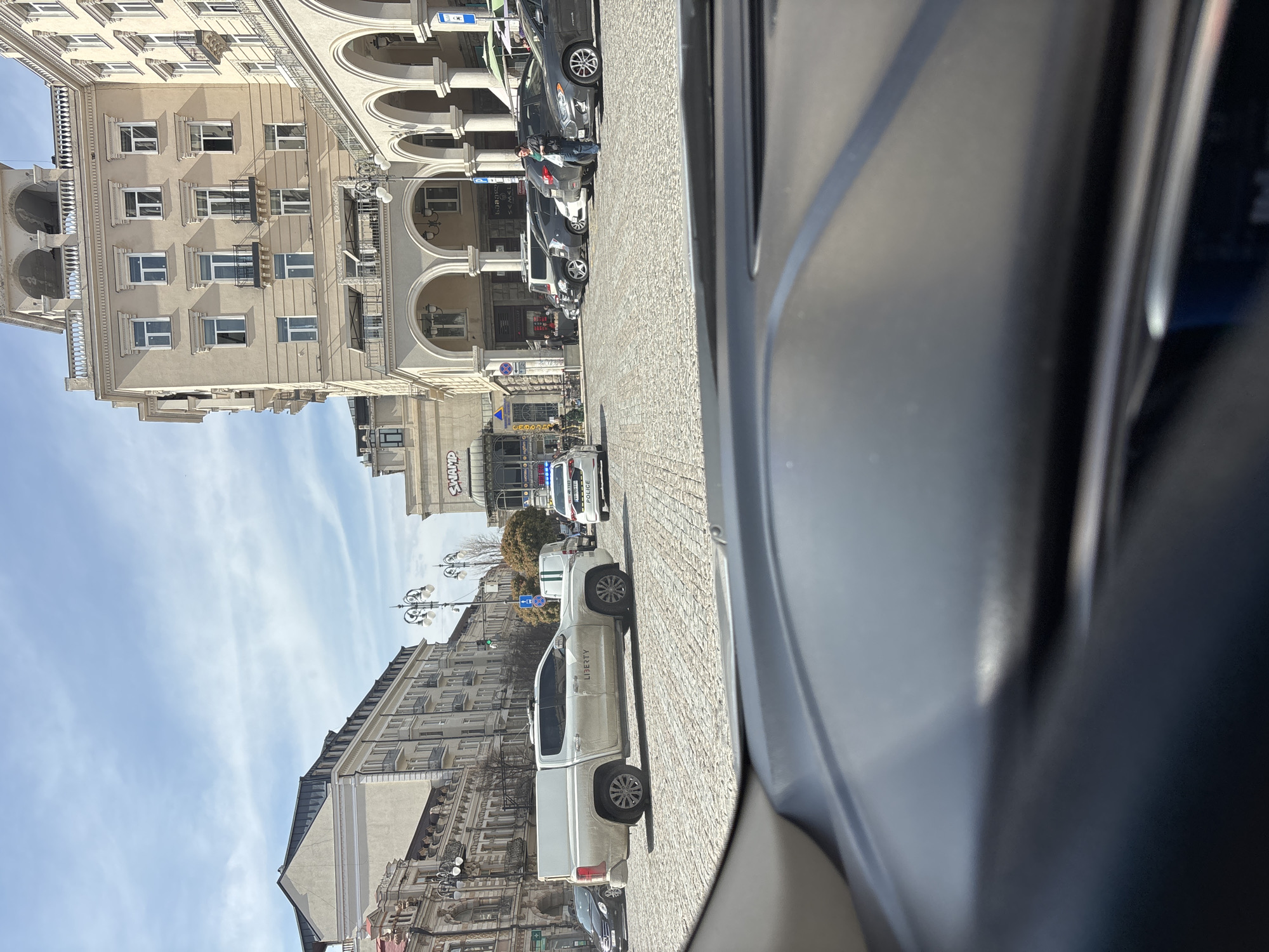The image shows a street scene in Tbilisi, Georgia, as viewed from inside a vehicle. The street is paved with cobblestones, and buildings line both sides of the street. The buildings are old, with architectural details, including arched colonnades, windows, and balconies. A white "LIBERTY" pickup truck is parked on the right side of the street. Further down the street, there are other parked cars, including a police car with flashing blue lights. A few people are standing near the colonnades of the building on the left. The sky is blue with scattered clouds. The overall impression is of a typical urban street scene in Tbilisi on a sunny day.