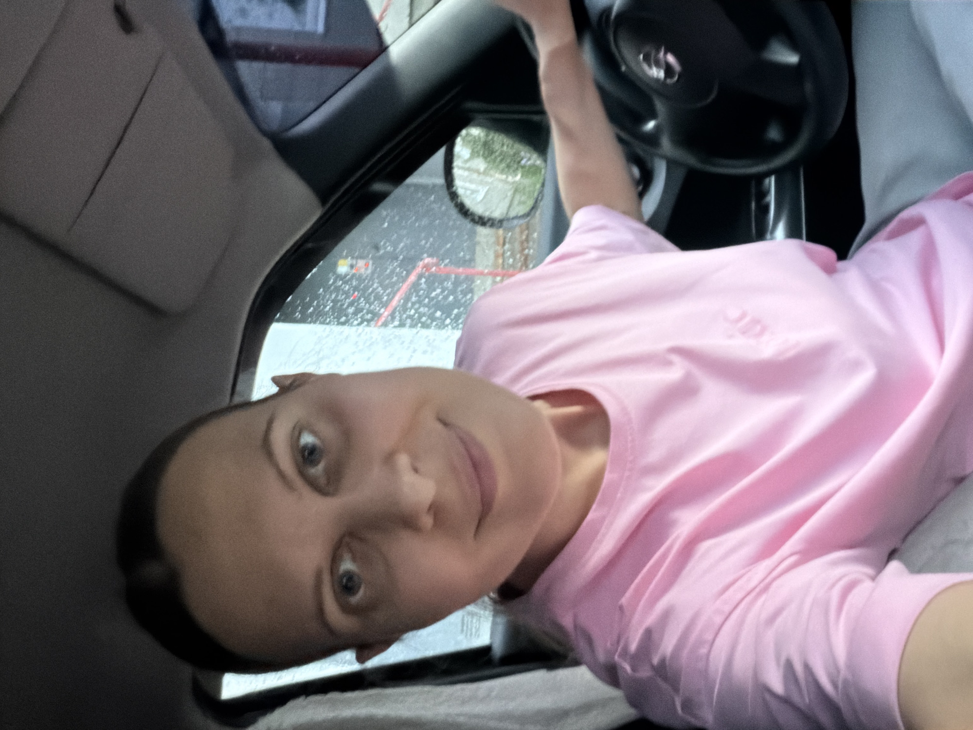 The image shows a woman sitting in the driver's seat of a car. She is wearing a pink t-shirt and looking at the camera, which is positioned in front of her. The car's interior is visible, including the steering wheel and part of the dashboard. Outside the car, it appears to be raining, with water droplets visible on the windshield and windows. A side mirror is also visible, reflecting a green, tree-lined area. The woman's expression is neutral. There is no visible text in the image. The scene suggests the woman is either waiting in her car or has just started driving. Given the context of Izhevsk, Russia, it could be any time of year, though the rain might suggest warmer months.