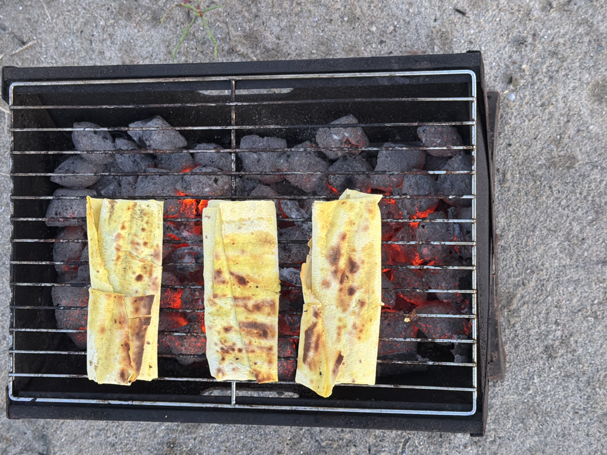 A close-up, overhead view shows three pieces of flatbread being grilled over glowing charcoal in a small, black metal barbecue. The bread appears to be yellow and slightly browned with char marks. The barbecue rests on a light gray, textured concrete surface. In the upper right portion of the image, a few dry green blades of grass are visible. The scene does not provide any information indicating the location in Odesa, Ukraine.