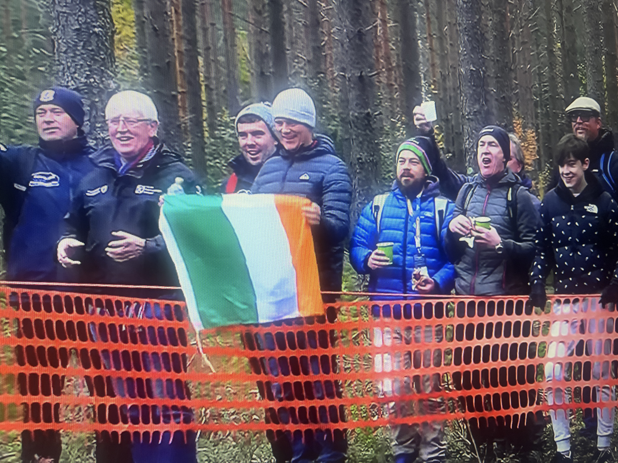 A group of people are gathered outdoors, with a forest in the background. A bright orange plastic mesh fence runs horizontally across the lower part of the frame. Several individuals are visible behind the fence. The person on the far left is wearing a dark blue jacket and a dark beanie with a logo. Next to them is a man with white hair wearing glasses and a dark jacket. Another man, wearing a grey beanie and a puffer jacket, is holding an Irish flag. To his right, a man in a blue puffer jacket with a green and black beanie is holding a cup. Further to the right, a person in a dark puffer jacket with a patterned design is visible. Behind them, a man wearing a cap is also present. The environment appears to be a wooded area, with trees forming the backdrop. There is no indication of the city of Masazir, Azerbaijan.