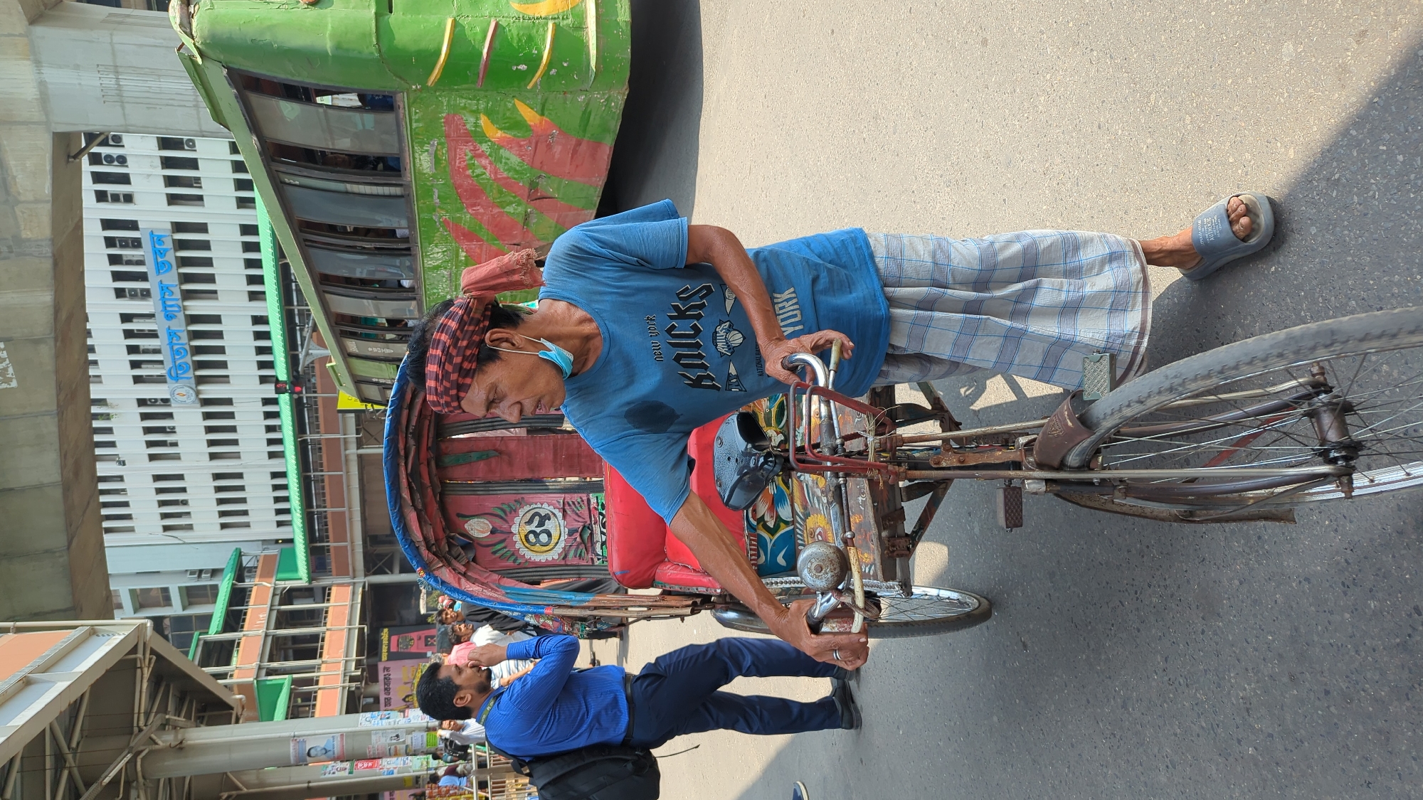 The image shows a man in Dhaka, Bangladesh, operating a rickshaw. The man is wearing a blue New York Knicks T-shirt, plaid pants, and blue sandals. He has a red and black checkered cloth on his head and a blue surgical mask hanging below his chin. The rickshaw is red with colorful decorations. The rickshaw operator is interacting with a man in a blue shirt and black pants standing in front of the vehicle. A green bus is visible behind the rickshaw, and a tall white building with dark windows stands in the background, appearing to read 'Titas Gas' in Bengali lettering. The scene is on an asphalt street.