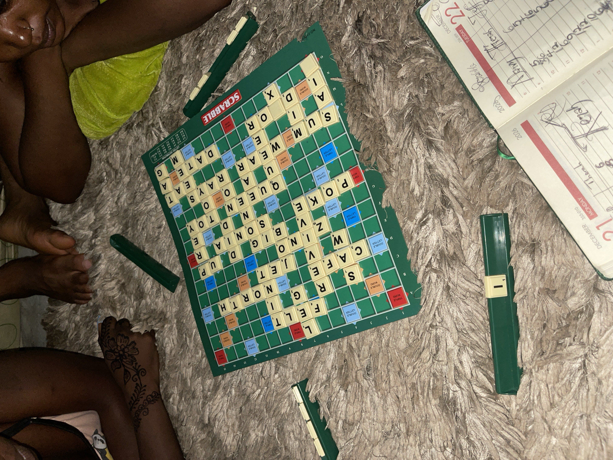This media file captures a casual indoor scene in Kuchingoro, Nigeria, likely in the evening or at night, as suggested by the artificial lighting. The primary activity is a game of Scrabble, laid out on a fluffy, light-colored rug or carpet.
At least three individuals are present, evidenced by visible body parts. One person's upper body is partially seen on the left, wearing a bright neon green garment, with dark skin. Another person's lower legs and feet are visible near the Scrabble board. A hand adorned with intricate henna art, featuring a floral design, is visible at the bottom of the frame, belonging to a third person.
The Scrabble board is mid-game, displaying numerous played words such as "FELT," "NORTH," "VJET," "SAFE," "POKE," "SUM," "WEAR," "QUEEN," "BINGONA," "DATE," "RAYS," "TOY," "SON," "UP," "OR," "X," "G," "M," "A," "I," "S," and "D." Several green Scrabble tile racks are placed around the board.
To the right, an open diary or planner is visible, showing entries for "DECEMBER 21 MONDAY 355-010" and "DECEMBER 22 TUESDAY 356-009," both dated "2026." Handwritten notes include "Scrabble," "Therah," "Rezah," and "Thank," along with a phone number "5042692206." The scene conveys a relaxed and engaged atmosphere of people enjoying a board game together.