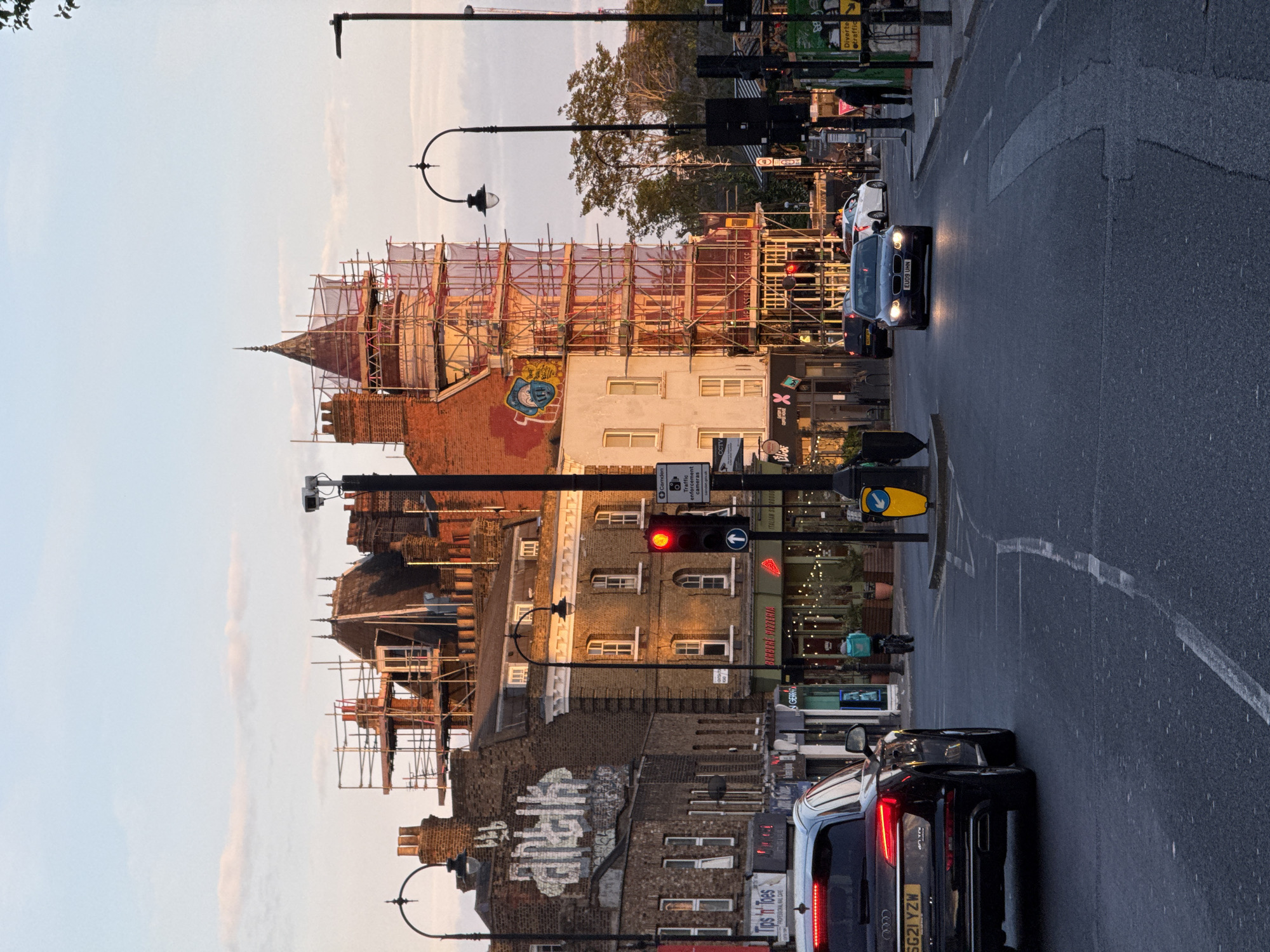 This street scene in London, United Kingdom, captures a mixture of residential and commercial buildings undergoing construction. The buildings are predominantly brick, with some covered in scaffolding, indicative of renovation work. Street lamps and traffic lights, one displaying a red signal, are visible along the roadside. Vehicles, including a black SUV with license plate SG21 YZW, and a dark car with license plate EU09 UHN, navigate the road. Graffiti is present on some buildings. Shops with signs like "Tips 'n' Toes" and "BURBERI PIZZERIA" indicate the commercial nature of the area. The presence of traffic enforcement cameras suggests the location is subject to monitoring.