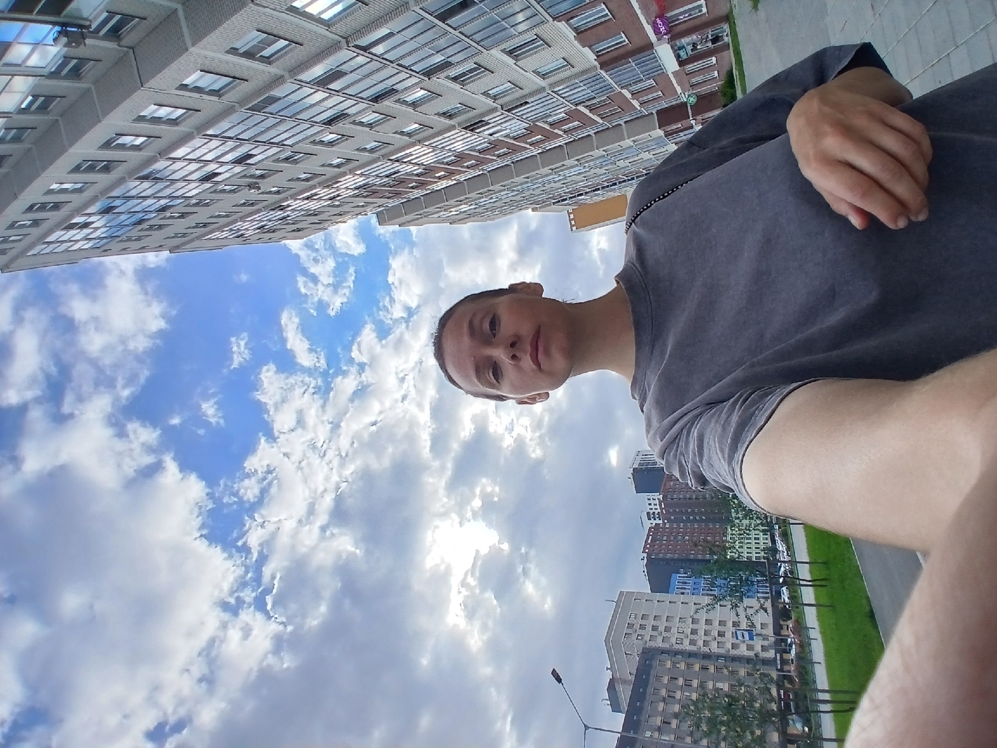 A medium-shot, low-angle, outdoor selfie taken by a person with light skin and dark hair pulled back, wearing a dark grey t-shirt, shows them standing on a paved walkway in front of modern, multi-story apartment buildings. The buildings are a mix of light grey and brick, with numerous windows and balconies, suggesting a new urban development. The sky above is a mix of blue sky and white clouds. In the background, to the left, is a street with parked cars and trees lining a grassy area. A lamppost is visible on the left side of the frame. The overall scene depicts a residential area in a city. Based on the architectural style and the presence of Russian signage, this location is identified as being in Yekaterinburg, Russia.