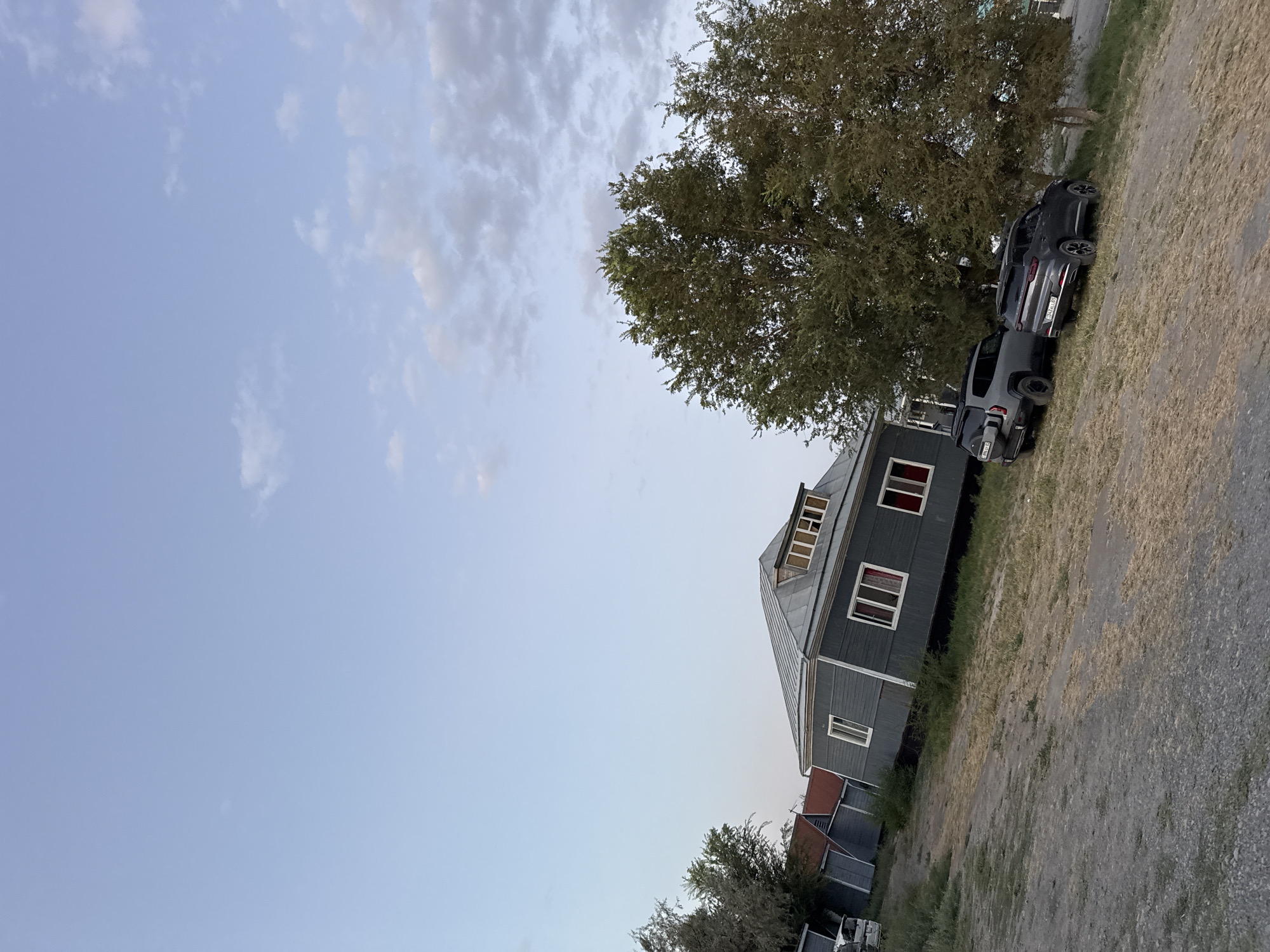 The image depicts an exterior view of several buildings with a natural landscape in the city of Urdzhar, Kazakhstan. In the foreground, a dirt and gravel path slopes upwards towards the right. Two dark-colored SUVs are parked on the grassy incline to the right of the path. Behind the SUVs, a large tree with green foliage dominates the scene. To the left of the tree and further back, a gray house with a silver metal roof and multiple windows is visible. This house appears to be older and has a slightly tilted appearance. Several other smaller structures, possibly sheds or outbuildings, are also present in the background, with some having red roofs. The sky above is a pale blue with scattered white clouds, suggesting it is either early morning or late afternoon. The overall scene is tranquil and rural.