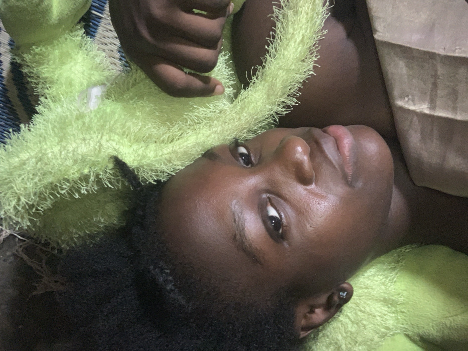 A forensic description of the scene depicts a close-up overhead view of a young individual, likely female, with dark skin and dark, braided hair, lying down. The subject's head is positioned towards the left of the frame, with her face turned slightly upwards, looking directly at the camera. Her expression is neutral. A small, dark stud earring is visible in her left earlobe.
A vibrant lime-green, highly textured, fuzzy fabric covers the right side of the subject and extends across the middle and upper right portions of the frame. A dark-skinned hand is visible on the right, resting on this green material. Portions of a blue and white patterned mat are discernible beneath the green fabric in the upper right background. A flat, brown, cardboard-like object is present in the lower right foreground. The overall lighting is subdued. This scene is observed in Agege, Nigeria.
