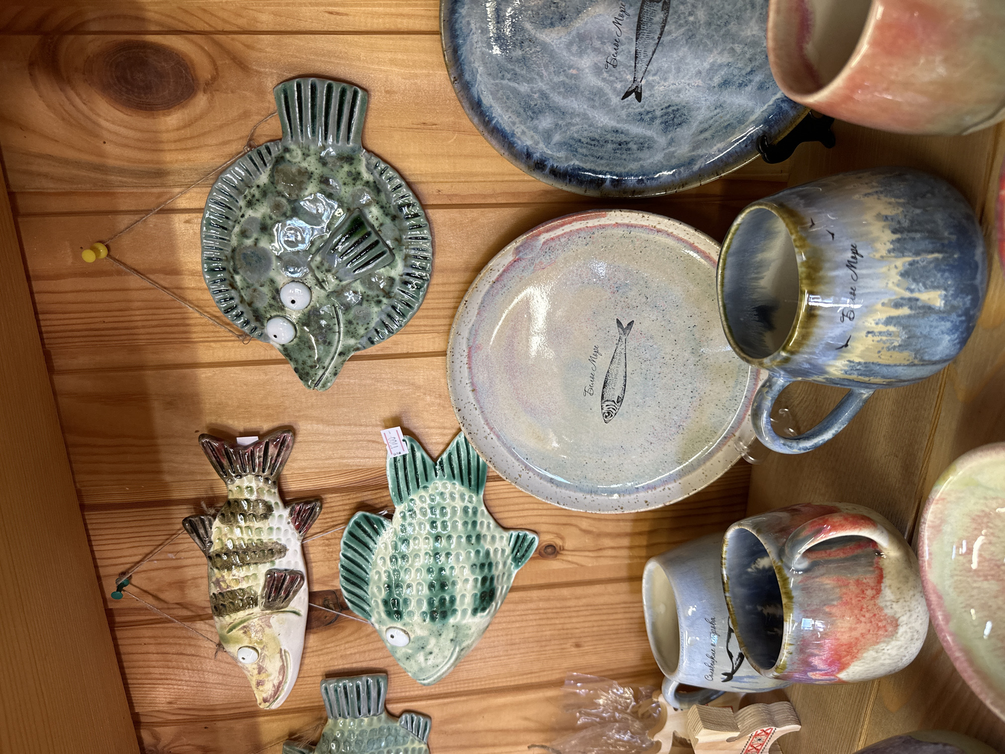 The image shows a close-up view of handcrafted ceramic items displayed on a wooden shelf. In the upper portion of the image, three ceramic fish decorations are hung on a wooden plank wall. The fish on the left is horizontally oriented with brown, green, and white stripes and a light brown belly. The middle fish is green with darker green speckles and large white eyes. The fish on the right is also green with a speckled texture and has darker green accents. Below the fish, two ceramic plates are visible. The plate on the left has a light cream-colored base with pinkish-red accents on the rim and a drawing of a fish with the inscription "Белое Море" (White Sea). The plate on the right is darker blue with a speckled pattern and a similar drawing of a fish with the same inscription. In the foreground, three ceramic mugs are visible. The mug on the left is light blue with an inscription and a drawing of birds. The middle mug has a rounded shape with a glaze that transitions from blue and grey at the top to brown and yellow at the bottom, also featuring the "Белое Море" inscription and birds. The mug on the right is a warm pinkish-orange color. The overall environment suggests a shop or display area for artisanal crafts, likely located in Solovetskiy, Russia, given the inscription on the plates and mugs.