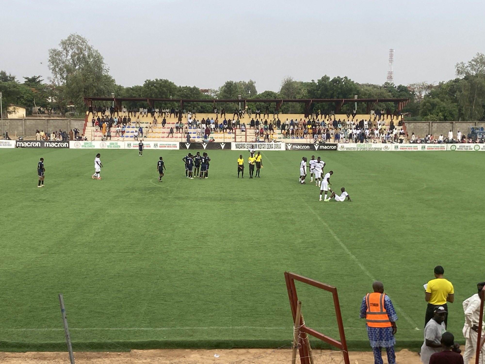 The image shows a soccer match in progress at Nasara Stadium Kofar Doka Zaria, likely in Tudun Wada, Nigeria. Two teams in contrasting uniforms are on the green field; one team wears dark blue and white, while the other wears white. Several referees in yellow and black stand among the players.
Spectators are seated in the stands behind the field. Banners with the logo "macron" and text mentioning "NASARA STADIUM KOFAR DOKA ZARIA," "KADUNA STATE MINISTRY OF SPORTS DEVELOPMENT," and "SEASON 2025/2026" are visible along the sidelines.
At the bottom right of the frame, individuals wearing vests are visible. A tower can be seen in the background behind the trees. A goalpost is partly visible at the bottom. The sky is overcast.