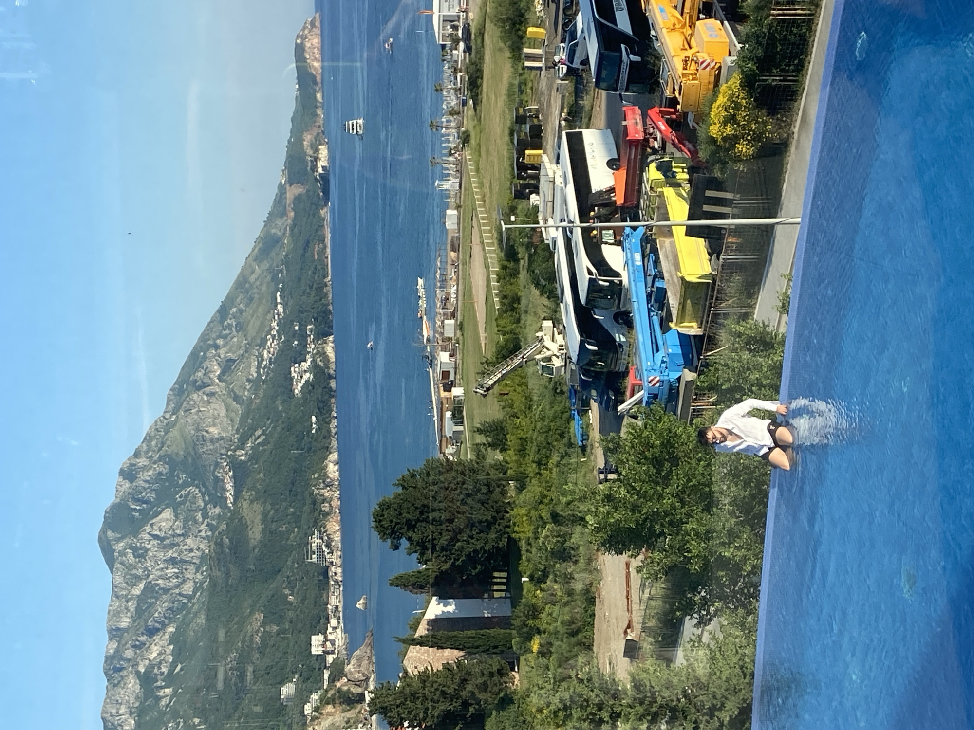 A medium shot depicts a man partially submerged in an outdoor, blue-tiled infinity pool. He is wearing a white long-sleeved shirt and dark-colored shorts. He is leaning forward slightly with his hands resting on the edge of the pool.
Behind the pool is a view of the coastline and a mountainous landscape, likely in Boreti, Montenegro. A body of water stretches from the shore towards the horizon. The mountains, covered in vegetation, dominate the background.
Closer to the camera, on the right side of the frame, are various construction vehicles and other equipment parked on a green, grassy area. A road runs through this area.
