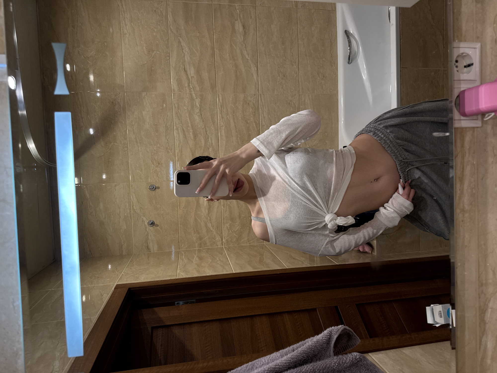 This media file shows a person taking a mirror selfie in a bathroom. The person is wearing a white semi-sheer long-sleeved shirt tied at the waist, exposing their midriff and a grey bra. They are also wearing grey sweatpants. The bathroom has beige tiled walls and a wooden vanity with a dark wood countertop. A white bathtub is visible to the right, along with a white electrical outlet and a pink object, possibly a hair dryer, plugged into it. The lighting appears to be artificial, suggesting it is likely indoors and not tied to a specific time of day. There are no clear indicators of weather conditions or strong emotions. The location is specified as Khimki, Russia, but there are no explicit visual cues within the image to confirm this. No visible text is present in the image.
