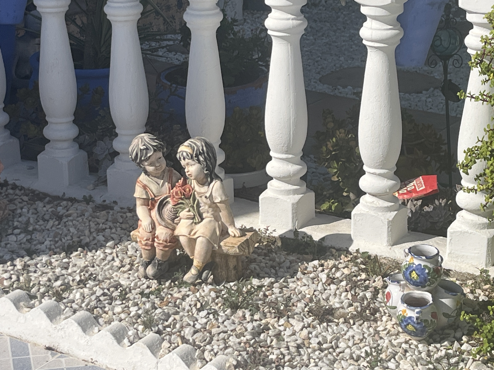 This is a forensic, matter-of-fact description of an outdoor scene.
The scene is a decorative garden area with white balustrades and planters. In the foreground, two small ceramic figurines of children are seated. The boy figurine, on the left, is wearing a peach-colored tunic and shorts. He is seated on the ground and appears to be looking at the girl figurine. The girl figurine, on the right, is wearing a peach-colored dress and a headband. She is seated on a small wooden stump and is holding a bouquet of red flowers. Her gaze is directed downwards.
To the right of the figurines, a cluster of decorative ceramic pots is visible. These pots are white with blue floral designs. Some are stacked. A bright red object, possibly a package or label, is visible behind the pots and in front of the balustrade.
The ground is covered in small, light-colored pebbles and gravel, with some sparse green vegetation growing through them. The balustrades are white and appear to be made of concrete or plaster, with decorative turned elements. Behind the balustrades, the environment suggests a garden setting with plants in blue planters. The lighting indicates it is daytime, with sunlight casting shadows.
While the image depicts a decorative garden setting, there is no specific visual information within the image that can definitively place it within the city of Torrevieja, Spain.