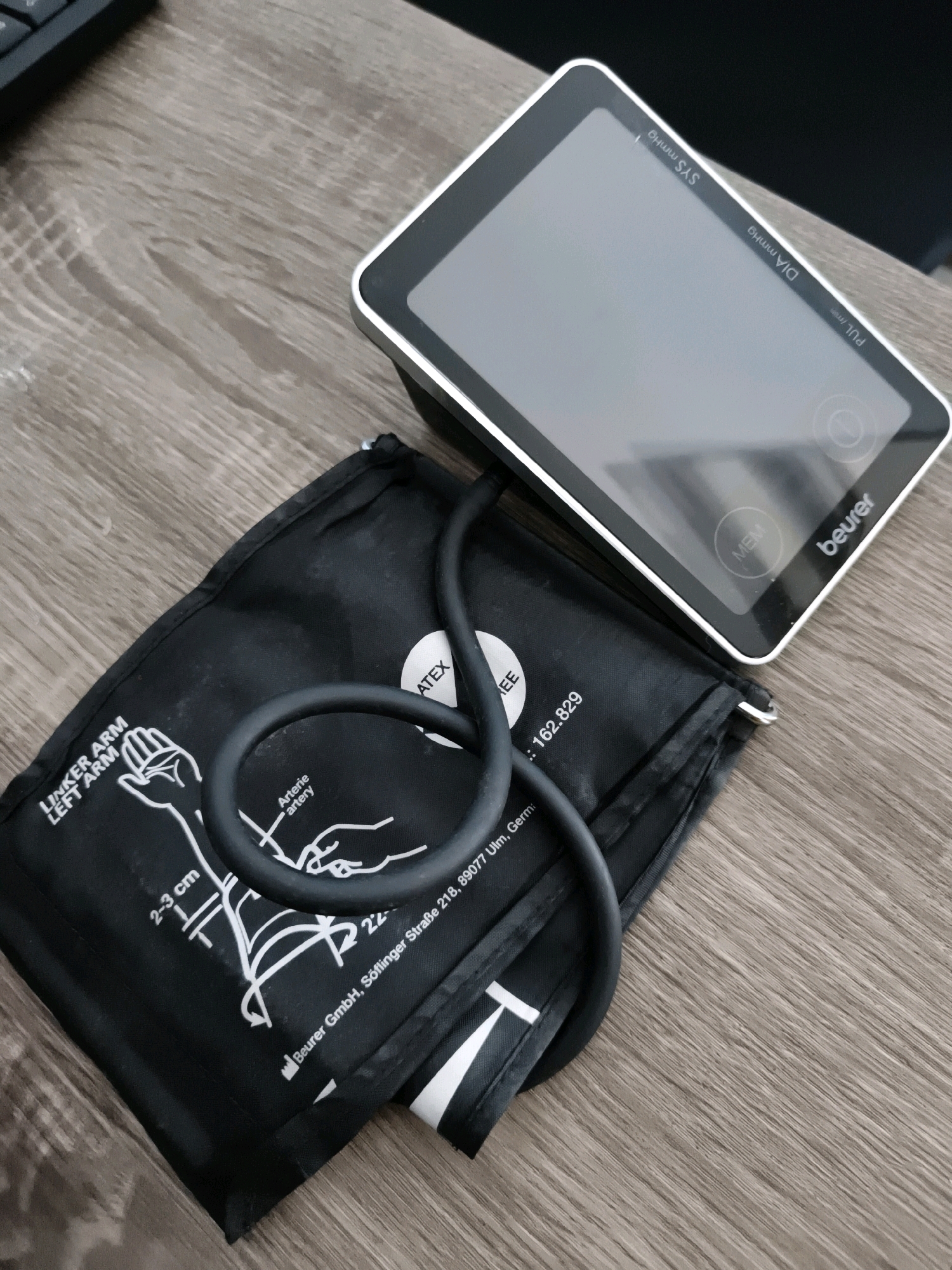 A Beurer brand blood pressure monitor is laid on a wooden desk, with its black cuff placed underneath the device. The monitor has a silver bezel and a dark grey touchscreen display. Text on the cuff indicates it is for the LEFT ARM and shows arm circumference guidelines from 2-3 cm. The device appears to be in a home or office environment. There are no people present in the image, and no discernible elements that place the image in Tombatu Dua, Indonesia.
