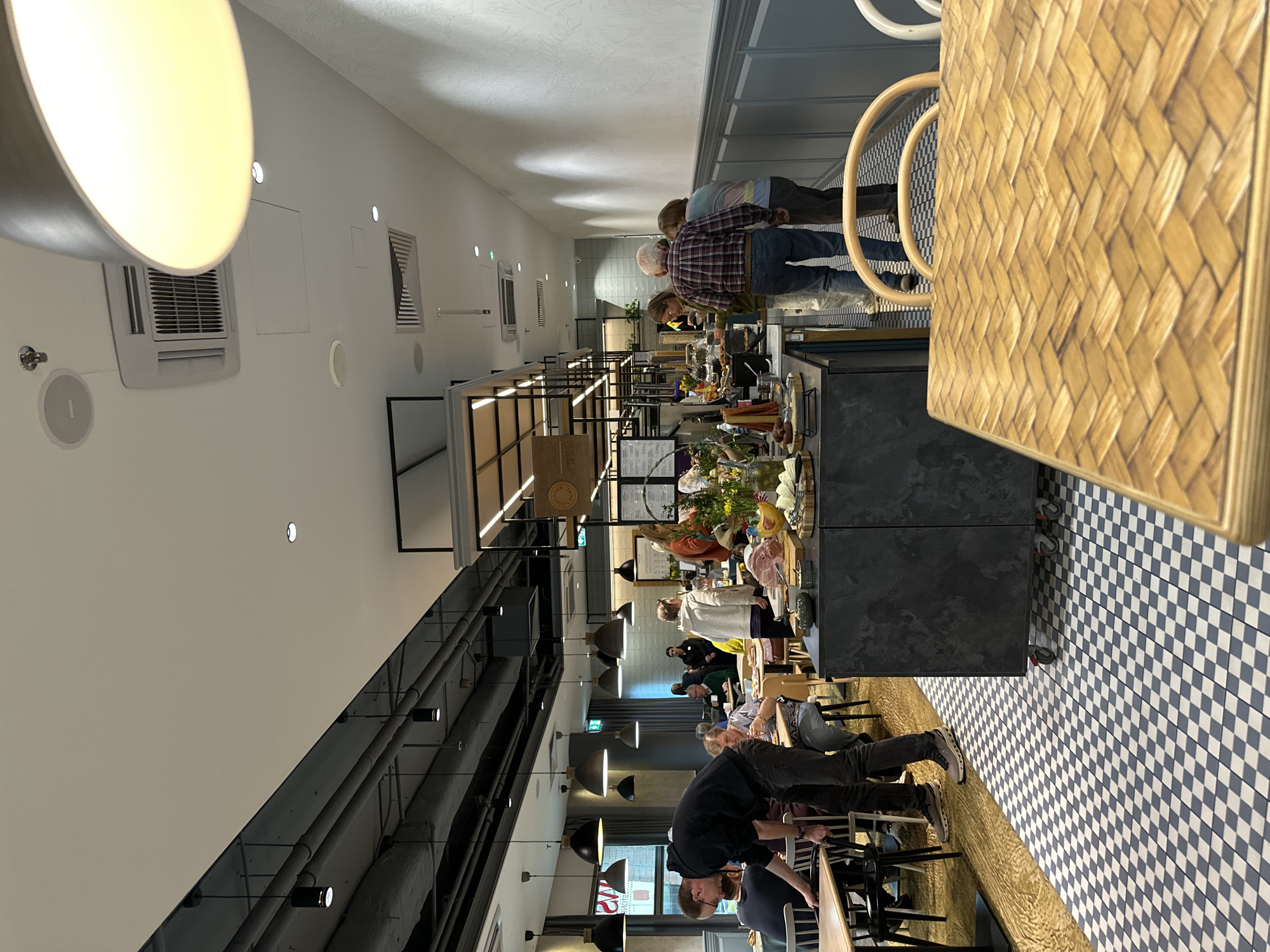 The image shows the interior of a restaurant in Wrocław, Poland. Customers are seated at tables and walking along a food counter. The restaurant has a modern design with black pipes on the ceiling, modern lighting fixtures, and a black and white checkered floor. The food counter features a variety of dishes. People are interacting with the food counter, apparently selecting dishes. Tables are made of light wood and placed next to a blue and white checkered floor. The restaurant appears to be in a well-lit urban environment.