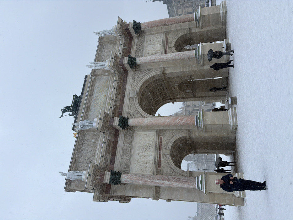 The Arc de Triomphe du Carrousel stands covered in snow on a winter day. Pedestrians walk nearby in the snow-covered ground. The sky is overcast and gray with heavy snow falling. The architecture of the arc is visible with its columns, sculptures, and intricate details. A person is walking in the foreground near the arc. The Louvre Pyramid is visible through one of the arches.