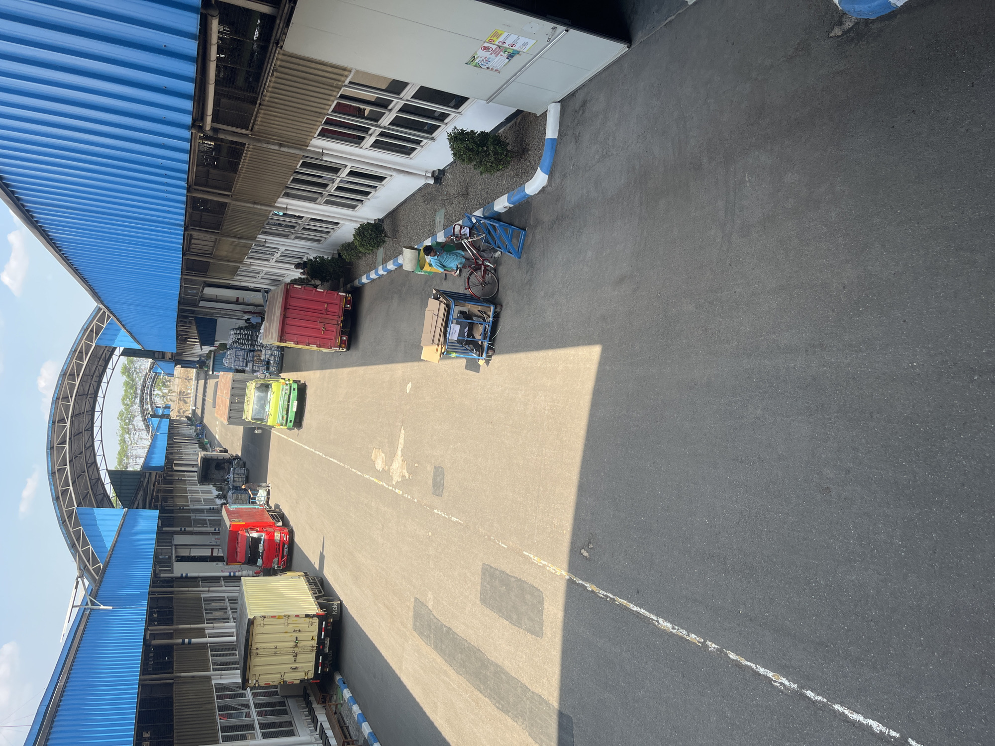 The image shows an outdoor industrial or warehouse area under a blue corrugated metal roof supported by a white metal frame. The area is paved with asphalt and marked with white lines. Several trucks are visible. On the left, a yellow box truck is parked. Further down the lane, a red box truck is parked, and a person wearing a blue shirt and shorts is standing next to a red bicycle and a trolley with cardboard boxes. To the right, a green truck is parked, and a red truck is in the background. Buildings with numerous windows line the right side of the area. The sky is clear and blue with some clouds. There are a few small green bushes planted along the edge of the buildings. The scene appears to be in an operational logistics or distribution center.