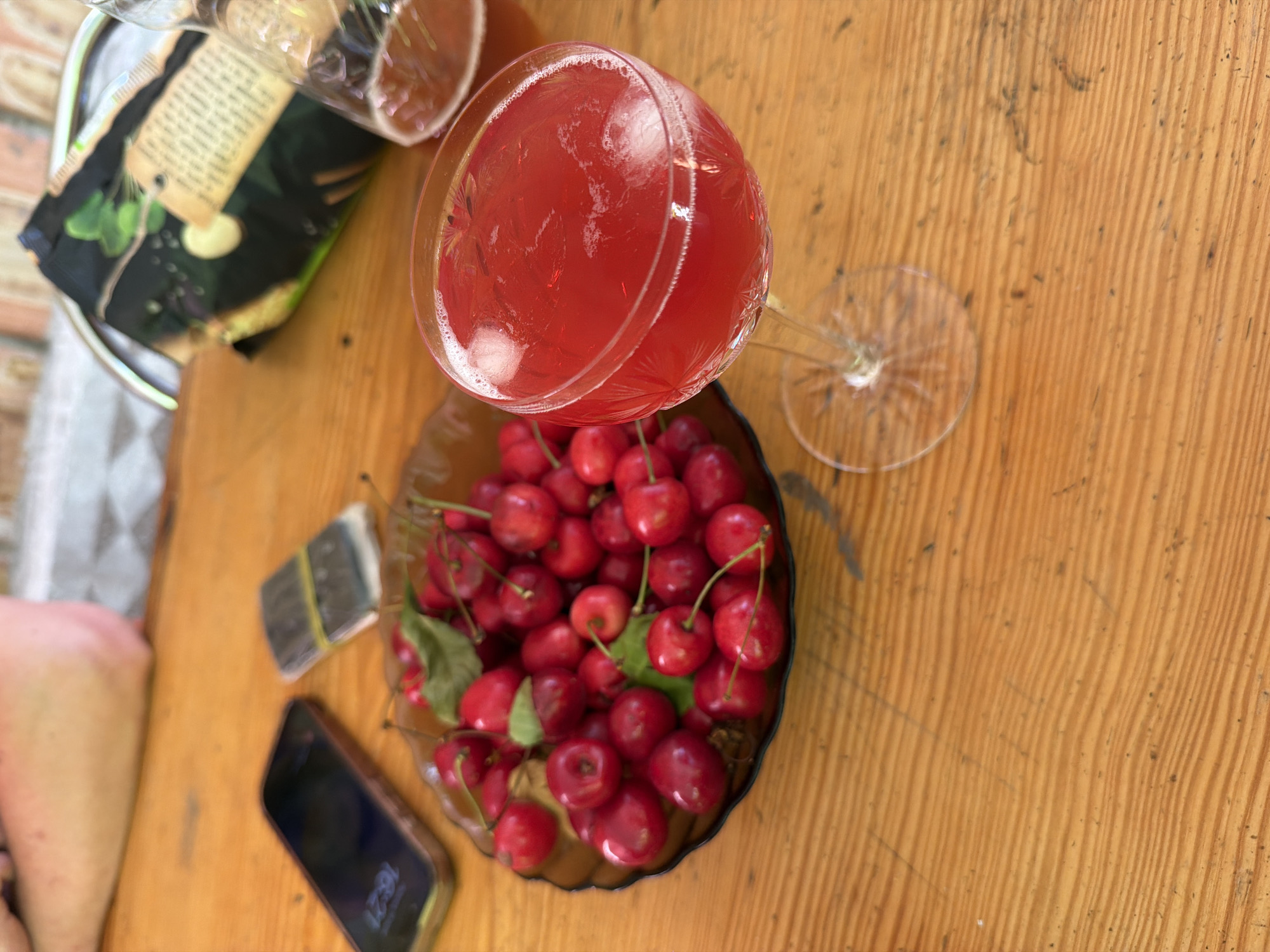 The image captures an overhead view of a wooden table, likely outdoors, with a collection of items suggesting a casual gathering. A stemmed glass filled with a vibrant red drink, possibly a cocktail or juice, sits prominently. Beside it, a dark bowl overflows with fresh, bright red cherries, some still attached to their stems and a few green leaves mixed in. To the left of the cherries, a black smartphone is visible, displaying the time "16:21". Next to the phone, a small stack of rectangular items, possibly packs of cigarettes or gum, is secured by a yellow rubber band. A person's leg, with visible skin and some freckles, is partially in the frame on the left side. In the background, a part of a black package with a rustic tag and green botanical imagery is also present. The lighting and warm tones of the wooden table suggest it might be daytime, possibly late afternoon, and the outdoor setting hints at pleasant weather. The overall impression is one of a relaxed, informal moment. No legible text is visible on the phone or other objects, other than potentially the branding on the black package, which is not clear enough to read. The scene is set in Obukhivka, Ukraine.