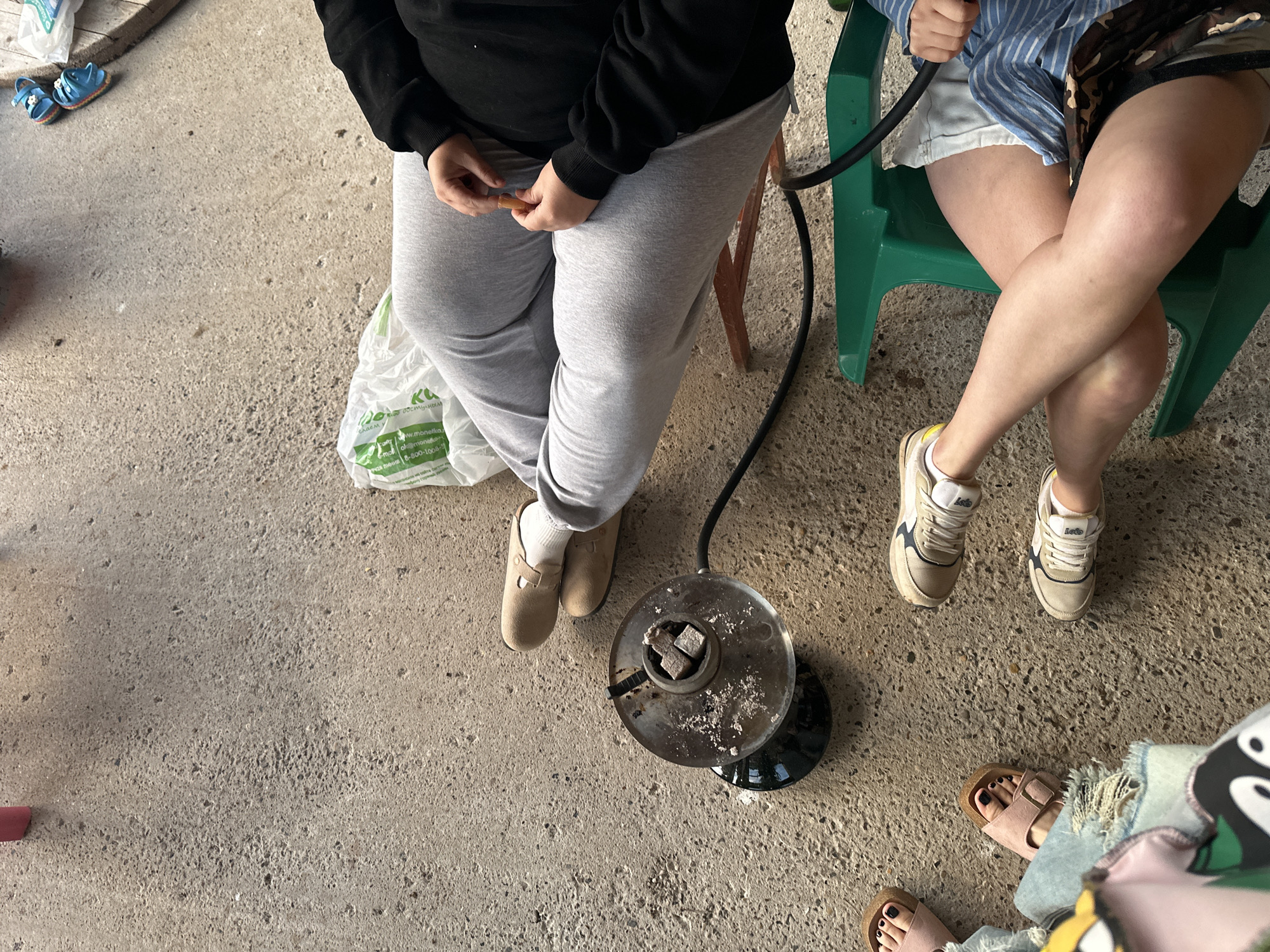 A high-angle shot shows a concrete patio area with several individuals present. In the foreground, a round, metal hookah bowl filled with charcoal sits on the ground, connected to a black hose. To the left, the feet of two individuals are visible; one person wears pink sandals with dark nail polish, and the other wears torn blue jeans. Further to the right, a person in light gray sweatpants and beige clogs is seated. Another individual, also in light gray sweatpants, is seen from the waist down, wearing a black hoodie and holding a small orange object. To the far right, a person is seated in a green chair, wearing light-colored shorts and a blue and white striped shirt, with their legs crossed. A white plastic bag rests on the ground near the seated individuals. The environment appears to be outdoors, possibly a backyard or patio. The location in the city of Пепеляева, Russia, is not evident from the visual information provided in the image.