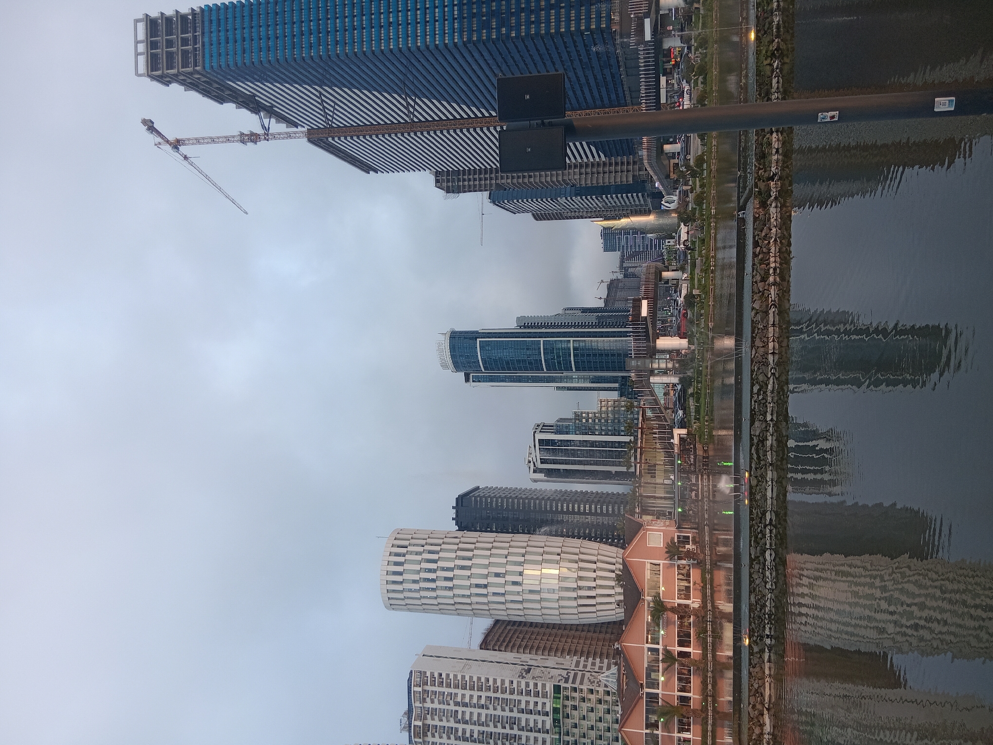 The image shows a skyline of buildings in Batumi, Georgia, reflected in the water. A tall, blue skyscraper is under construction, with a crane visible near its top. Several other high-rise buildings, including one with a cylindrical, white patterned design, line the waterfront. A concrete walkway with a stone edge separates the buildings from the water. A pole with lights or sensors is positioned prominently in the foreground, partially obscuring the buildings. The sky is overcast.
