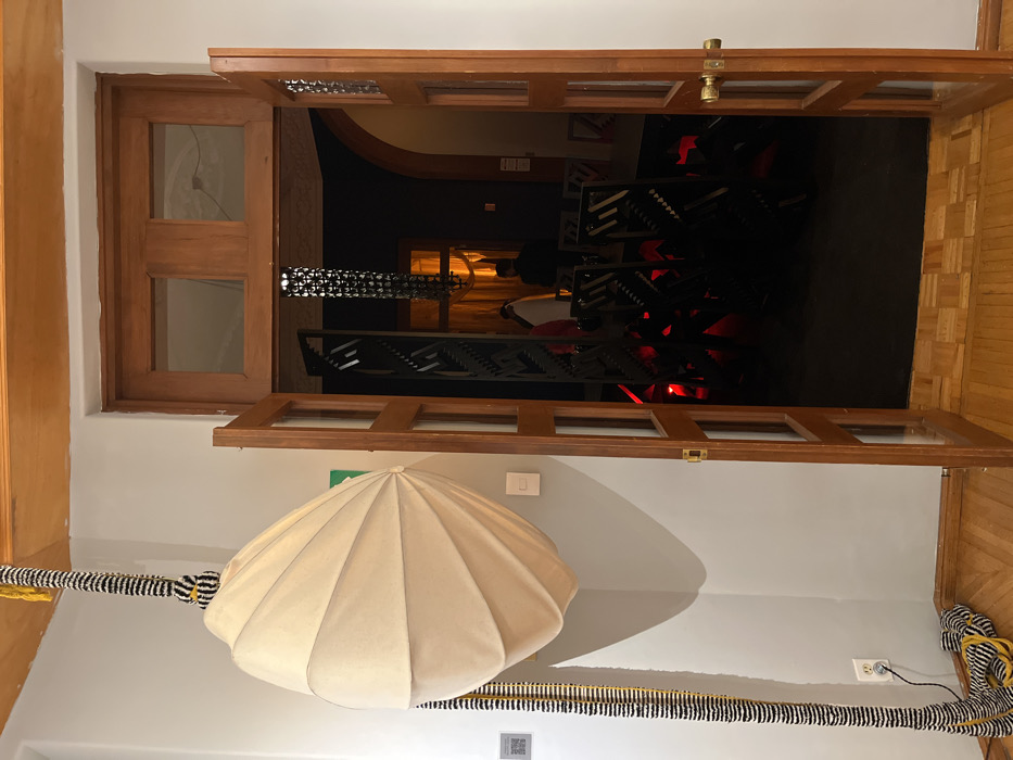An open, brown-framed glass door reveals a dark interior with an abstract black metal structure illuminated by red lights. A white umbrella-shaped object hangs on the light gray wall in the foreground, with a black and yellow striped cable extending across the bottom. A wood-paneled floor is partially visible on the right. The door's wooden frame matches the floor's texture and color. A light switch and a small QR code are visible on the wall.