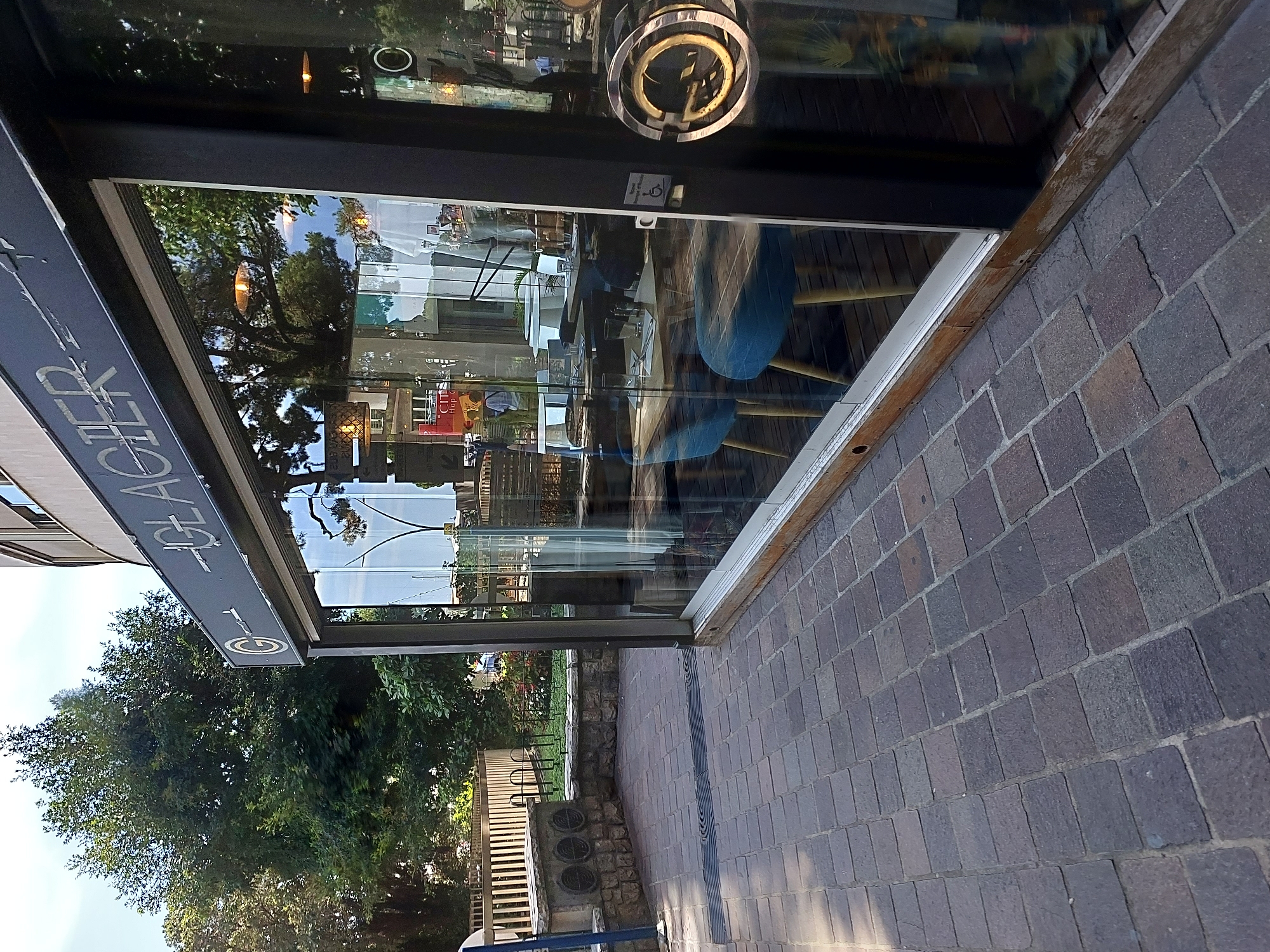 The image shows a storefront with a sign that reads "GLACIER". The storefront has large glass windows that reflect the trees and sky. Inside the store, tables and chairs are visible, suggesting it is a restaurant or cafe. The pavement outside is made of brick. A decorative metal handle is on the glass door. The location is the city of Monaco, Monaco.
