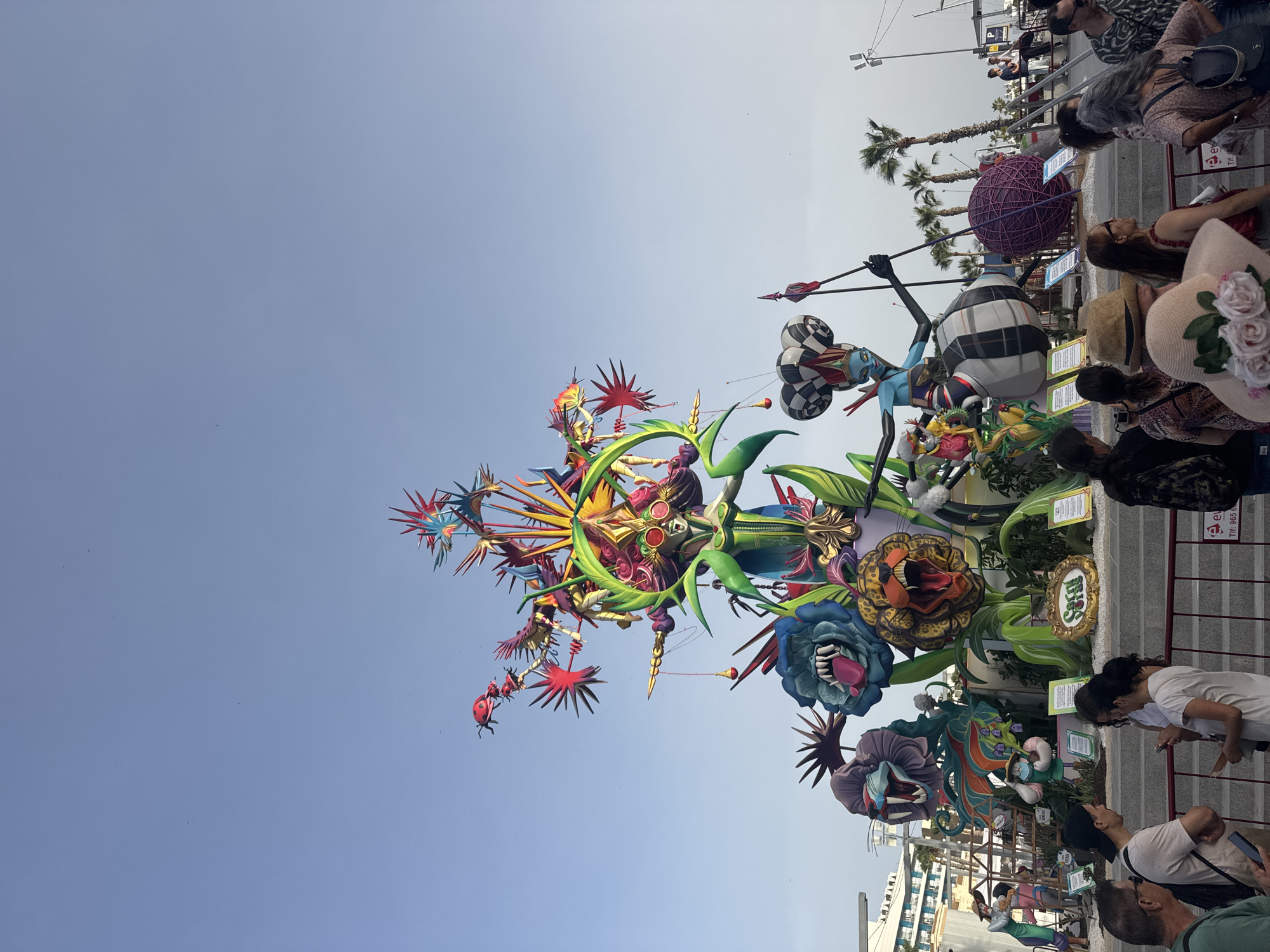 The image depicts a large, colorful monument, likely a "ninot" from the Fallas festival, in Alicante, Spain. The monument is elaborate, featuring fantastical figures, large animalistic heads with open mouths, and vibrant floral elements. A central female figure with blue skin, a jester-like hat, and elaborate clothing stands astride a large striped sphere. Smaller figures and mythical creatures are integrated into the structure. The monument is surrounded by a crowd of people, some of whom are looking at the monument, while others are engaged in conversation or looking at their phones. Palm trees and buildings are visible in the background, suggesting an outdoor urban setting. The sky is clear and blue. A low barrier with signage is in the foreground, indicating a viewing area or event space.