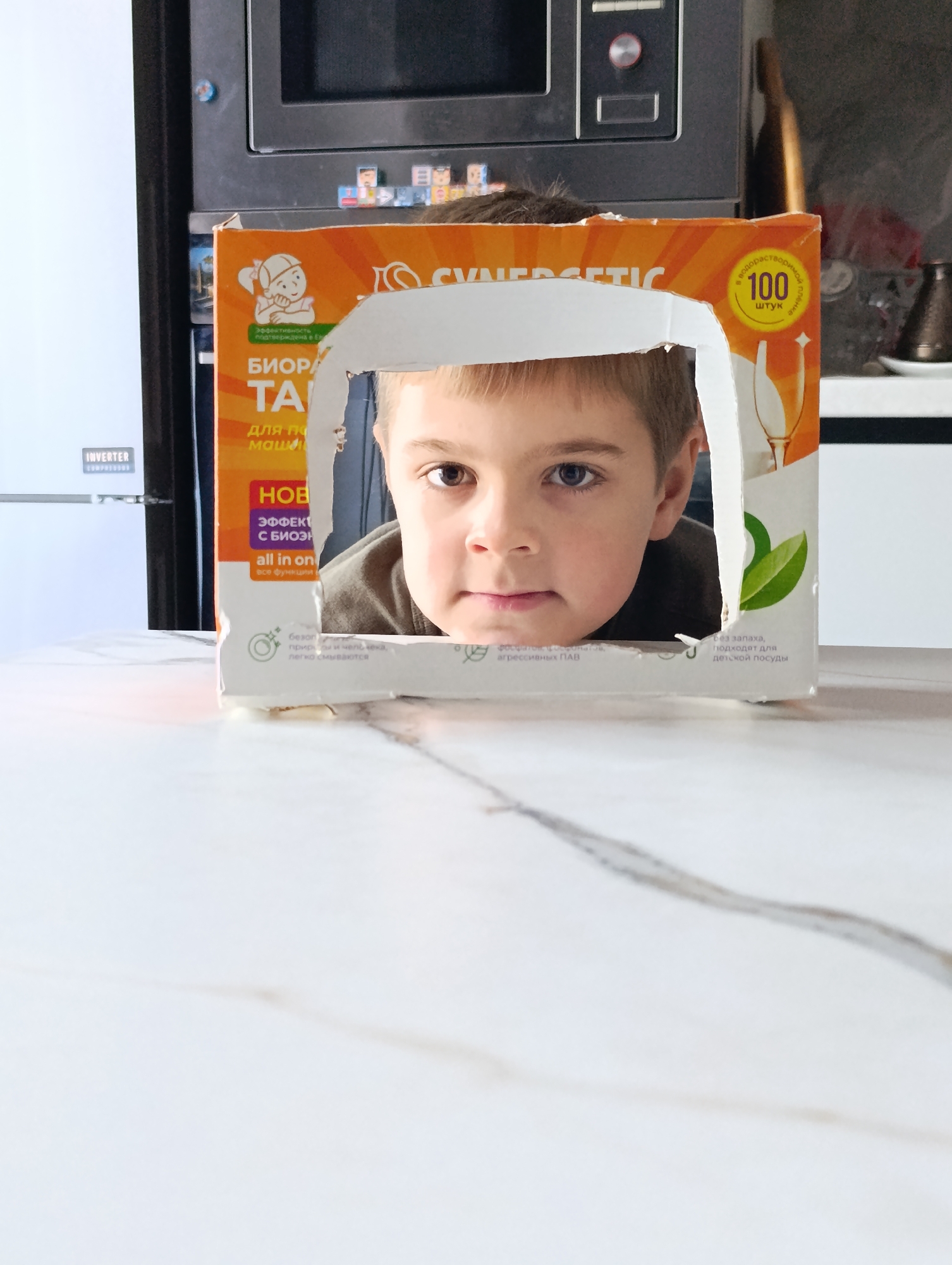 A young boy is peering through a hole cut in a cardboard box. The box has Russian text and images printed on it, indicating it likely contained a product. The boy's face is the main focus, framed by the makeshift cutout. The box is resting on a white countertop, with a refrigerator and microwave visible in the blurred background. The scene is indoors, presumably in a kitchen setting. The brand "SYNERGETIC" is printed on the box. The text on the box includes references to "БИОРА", "EFFECT С БИОЭН", "all in one", "100 штук" and "Эффективность подтверждена в Ел". There is no city depicted in this image.