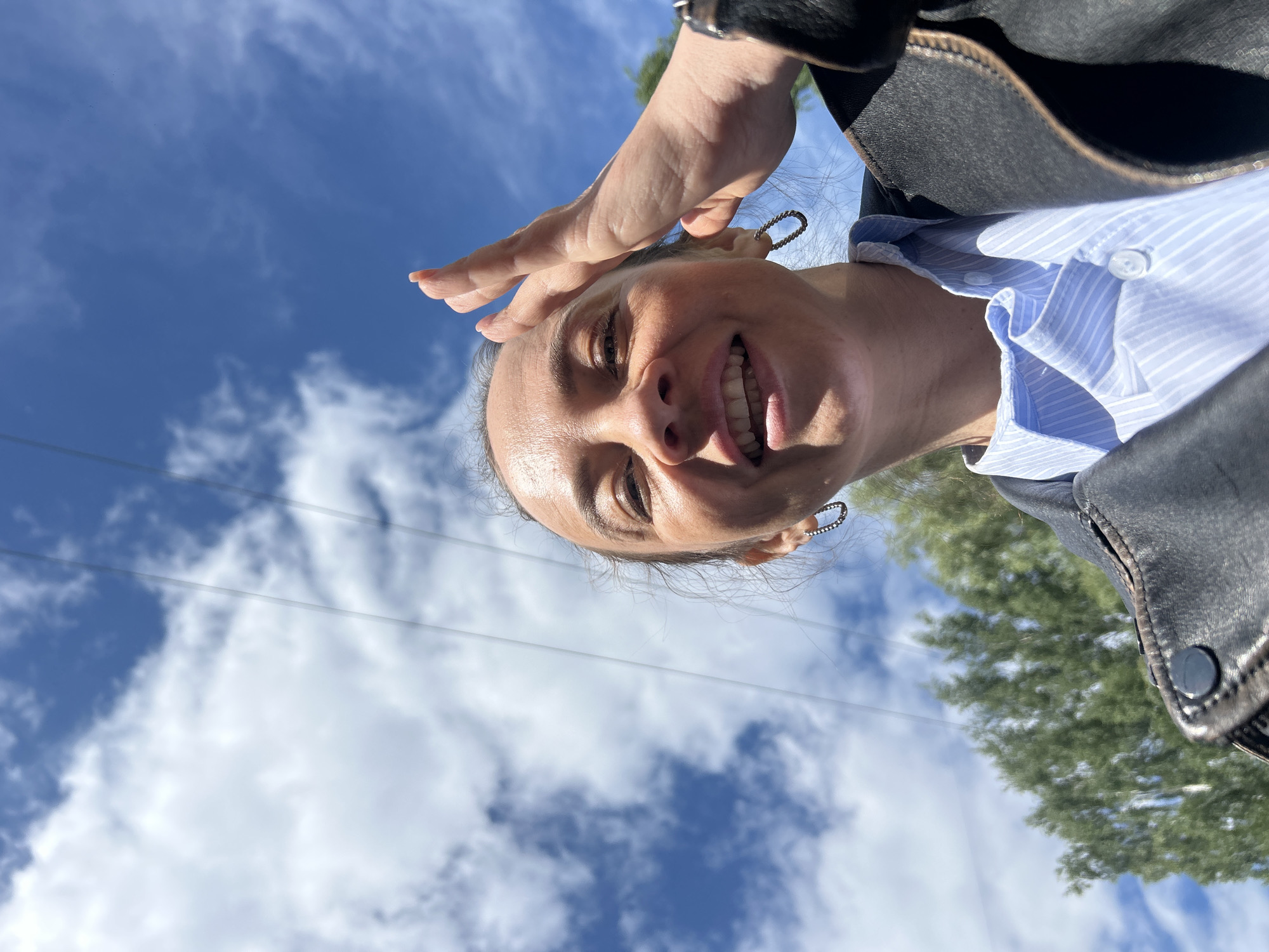 A woman is photographed from a low angle, looking down at the camera. She is smiling and partially shielding her eyes with her left hand. She is wearing a light blue, striped shirt, and a black leather jacket. The background consists of a partly cloudy blue sky. Power lines are visible. Based on the presence of Russian power lines, trees, and the woman's appearance, it is plausible that this image was taken in Chaikovsky, Russia.