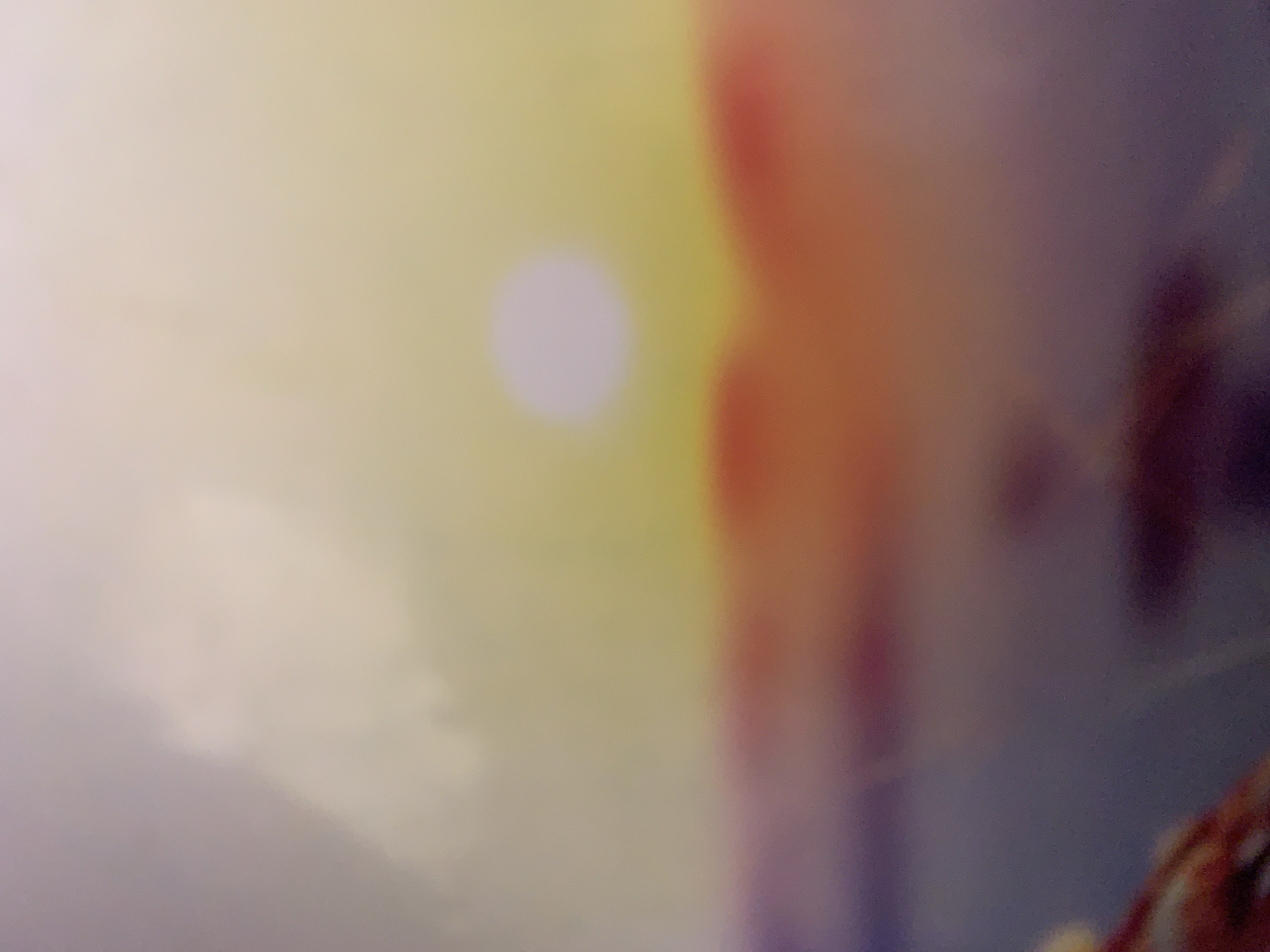 This is a highly abstract, blurry, and overexposed image with a predominantly soft yellow and pink hue. In the center-left, a pale, circular light source is visible, possibly representing the sun or a lightbulb. To the right, there are indistinct shapes and color gradients of orange, purple, and dark brown. The overall impression is one of extreme blurriness, with no clear subjects, objects, or activities discernible. The image does not provide any information about the time of day, weather, location cues, emotions, or visible text.