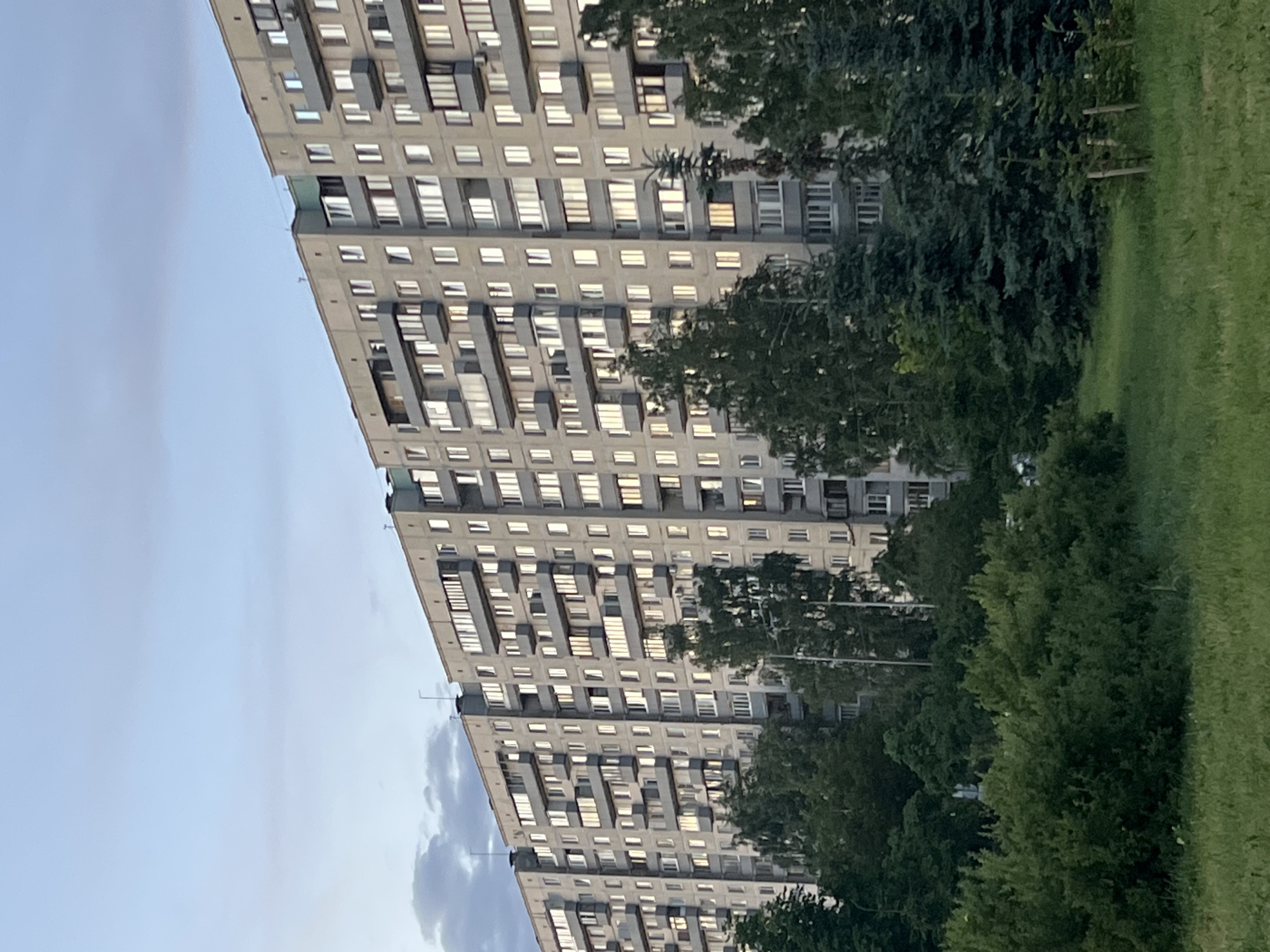 The image shows a large, multi-story apartment building in Ladozhskoe Ozero, Russia. The building is constructed of concrete panels and has a uniform design with many windows and balconies. The facade is a light grey color, and the balconies have railings. The building is surrounded by lush green trees and grass, suggesting a park-like or residential area. The sky is a pale blue with some scattered clouds. The overall impression is of a typical urban residential landscape. There are no people visible in the image.